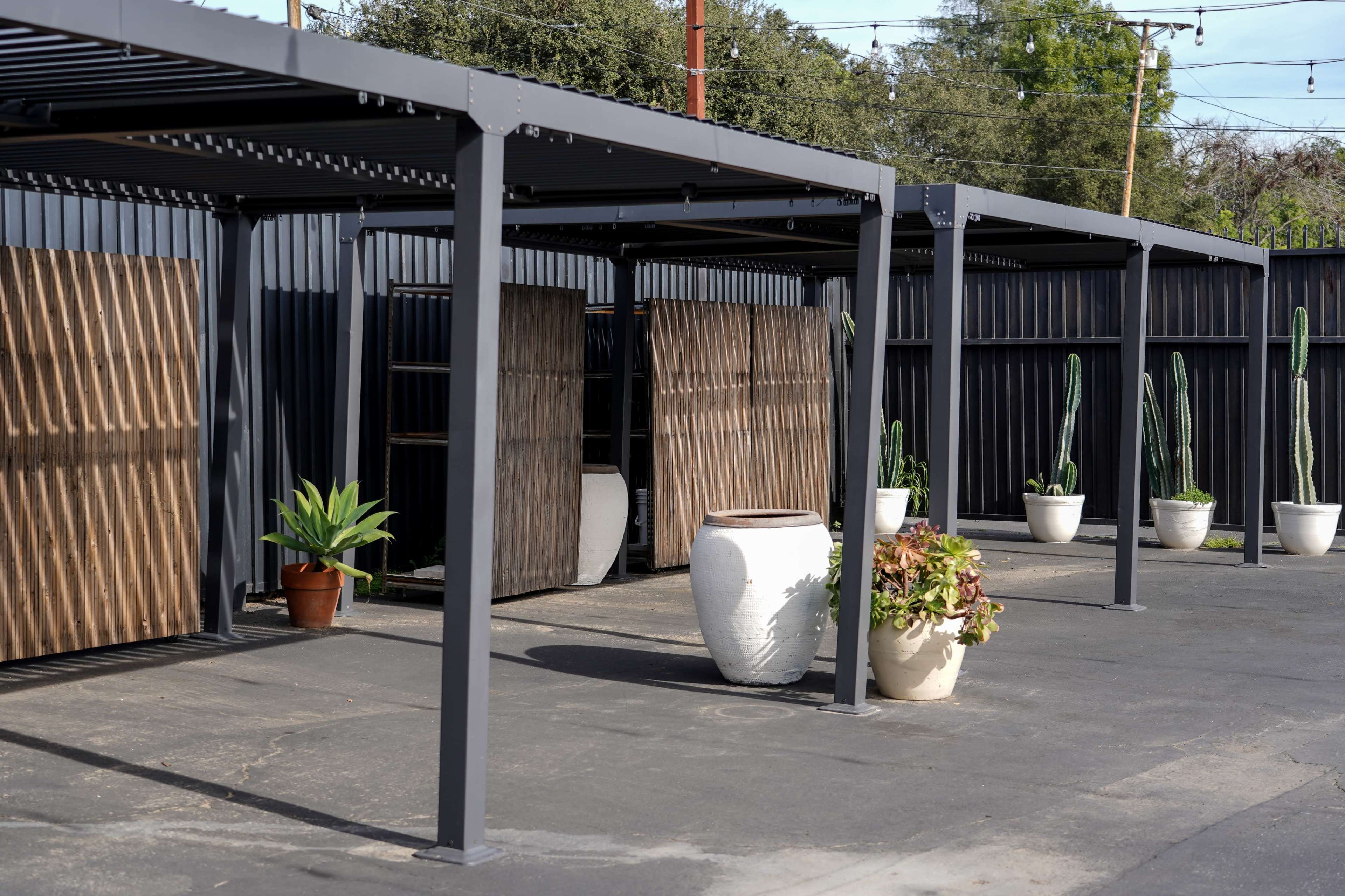 The image shows a modern outdoor area with metal structures, potted plants, and wooden panels set against a black fence.