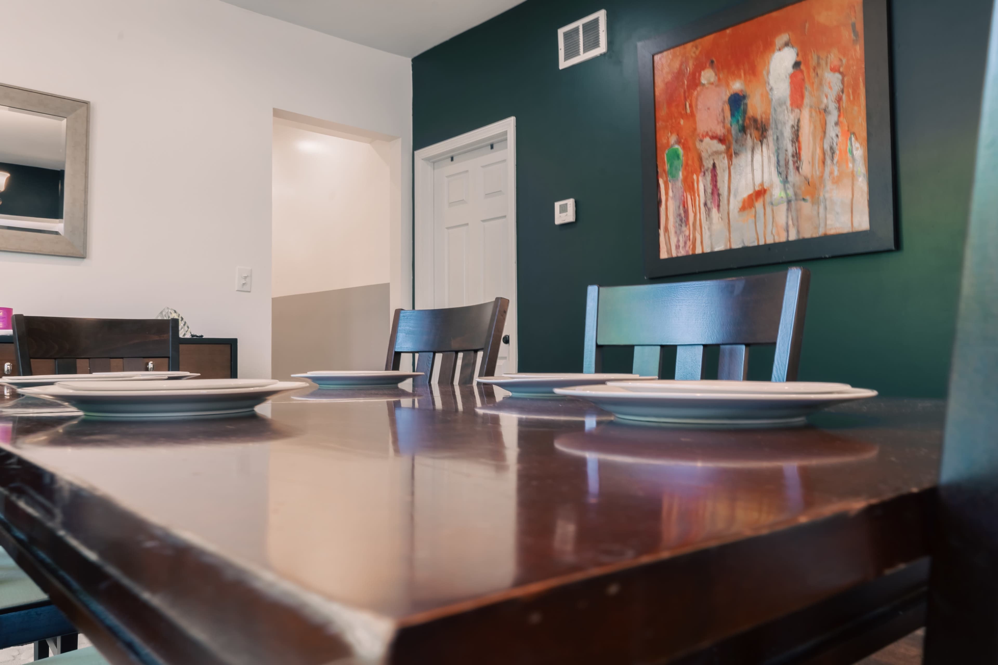 A dining table set with plates in a room featuring dark green walls and an abstract painting.