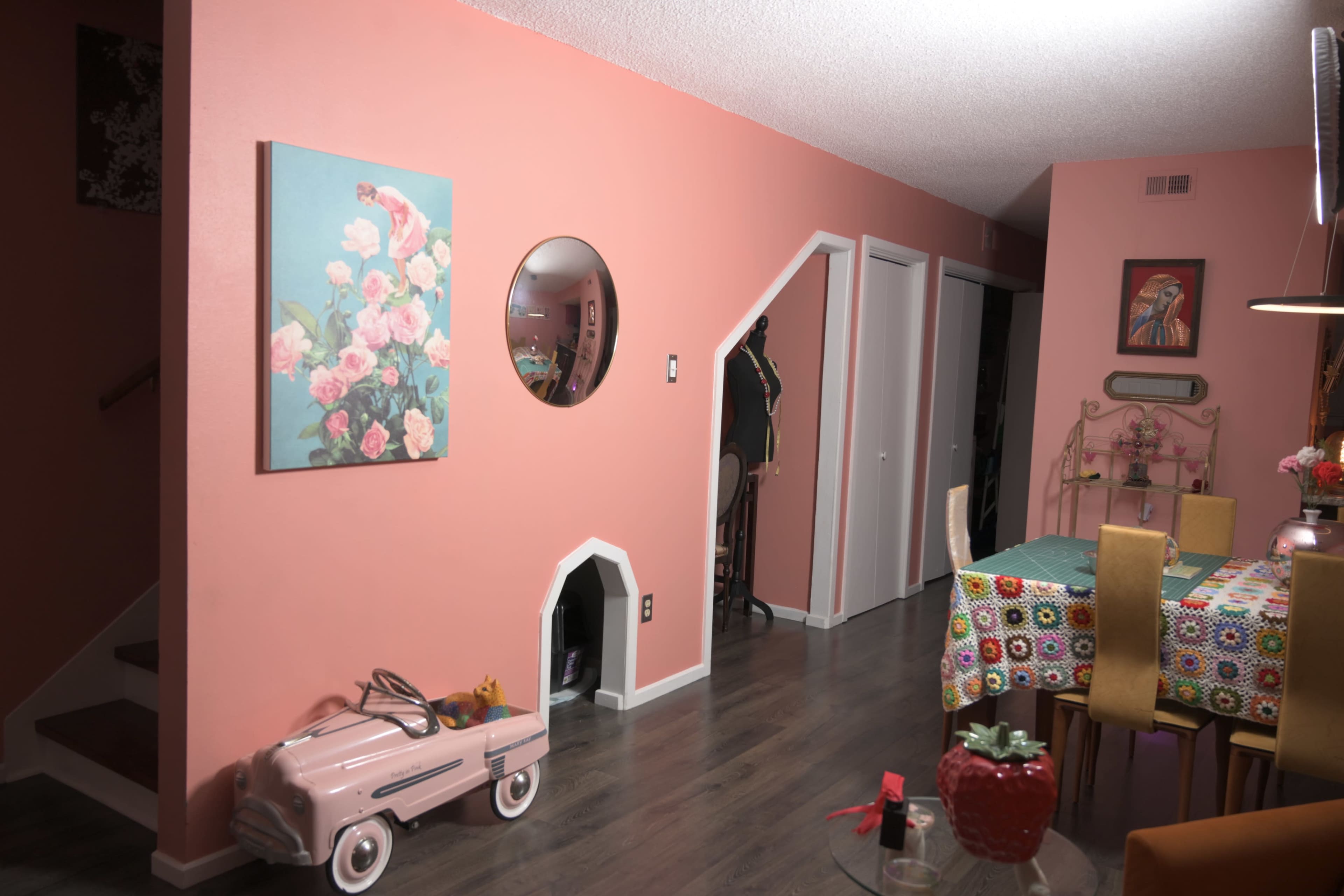 The room features a pink wall with a floral painting, a round mirror, a small toy car, and a dining table covered with a colorful tablecloth.