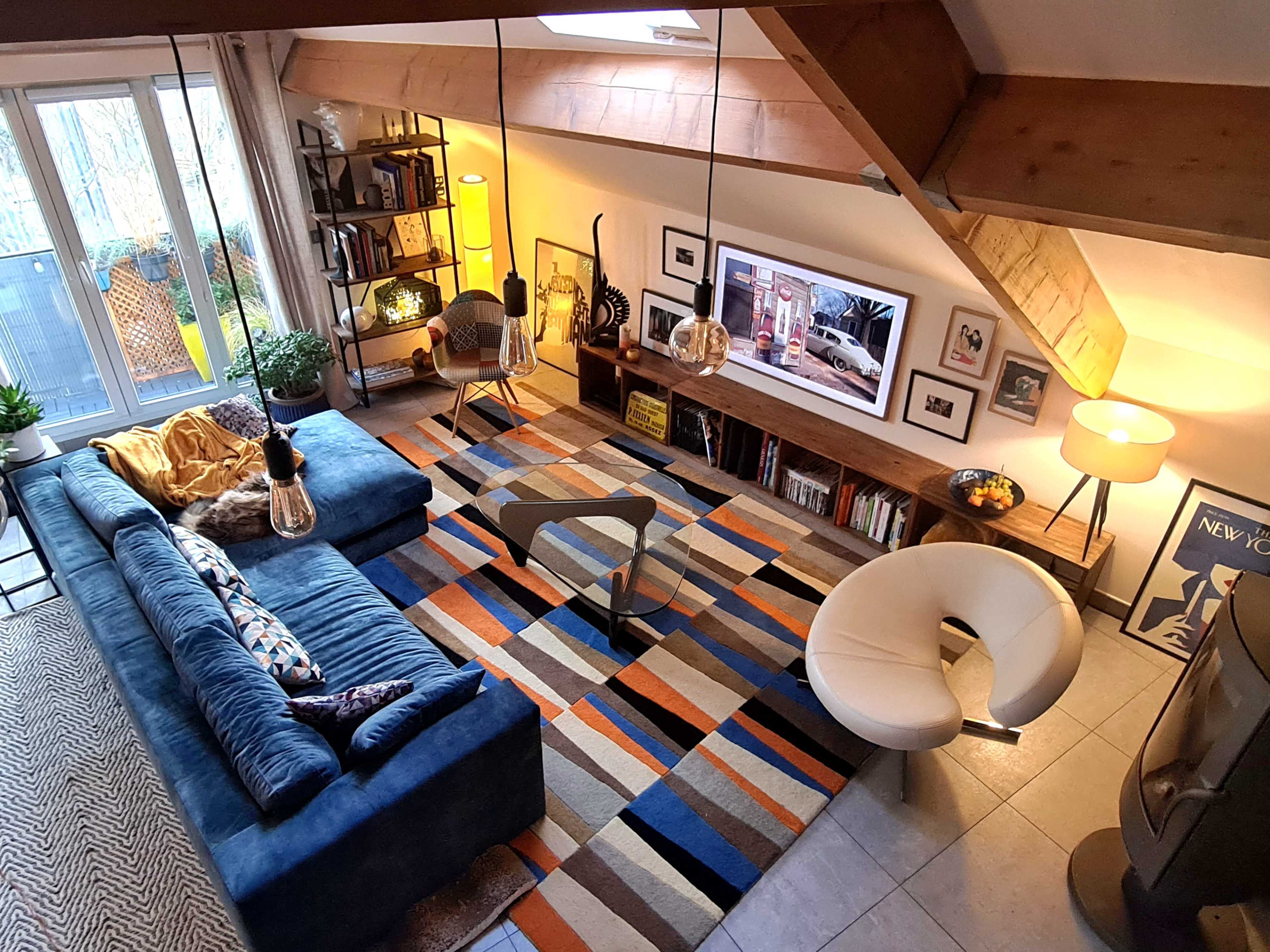 A stylish living room featuring a blue sectional sofa, a round white chair, a patterned area rug, and a wall-mounted television above a wooden shelf, with large windows allowing natural light to enter.