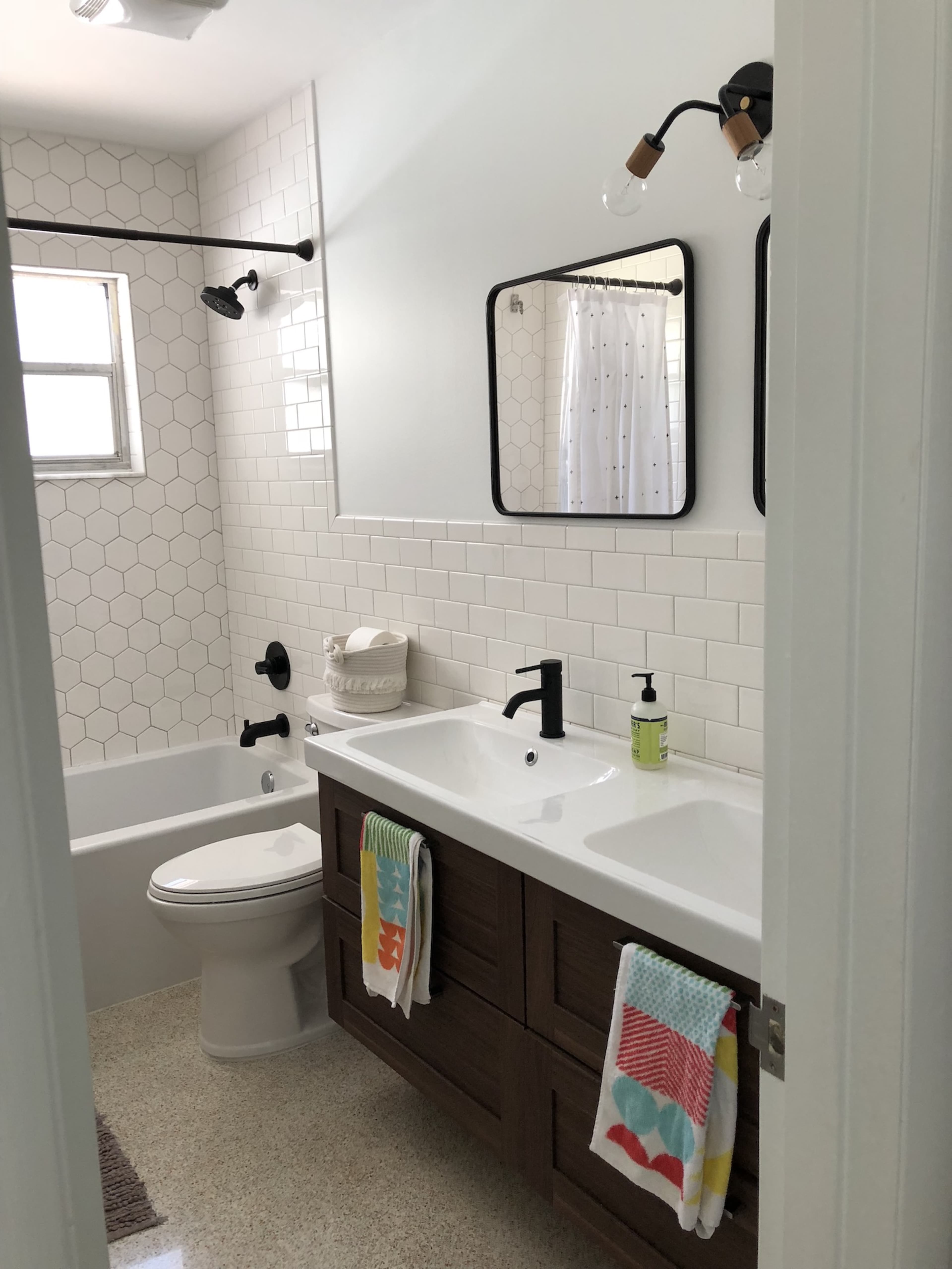 A modern bathroom features a white tiled wall, a double sink vanity with wooden cabinets, a bathtub, and a window letting in natural light.