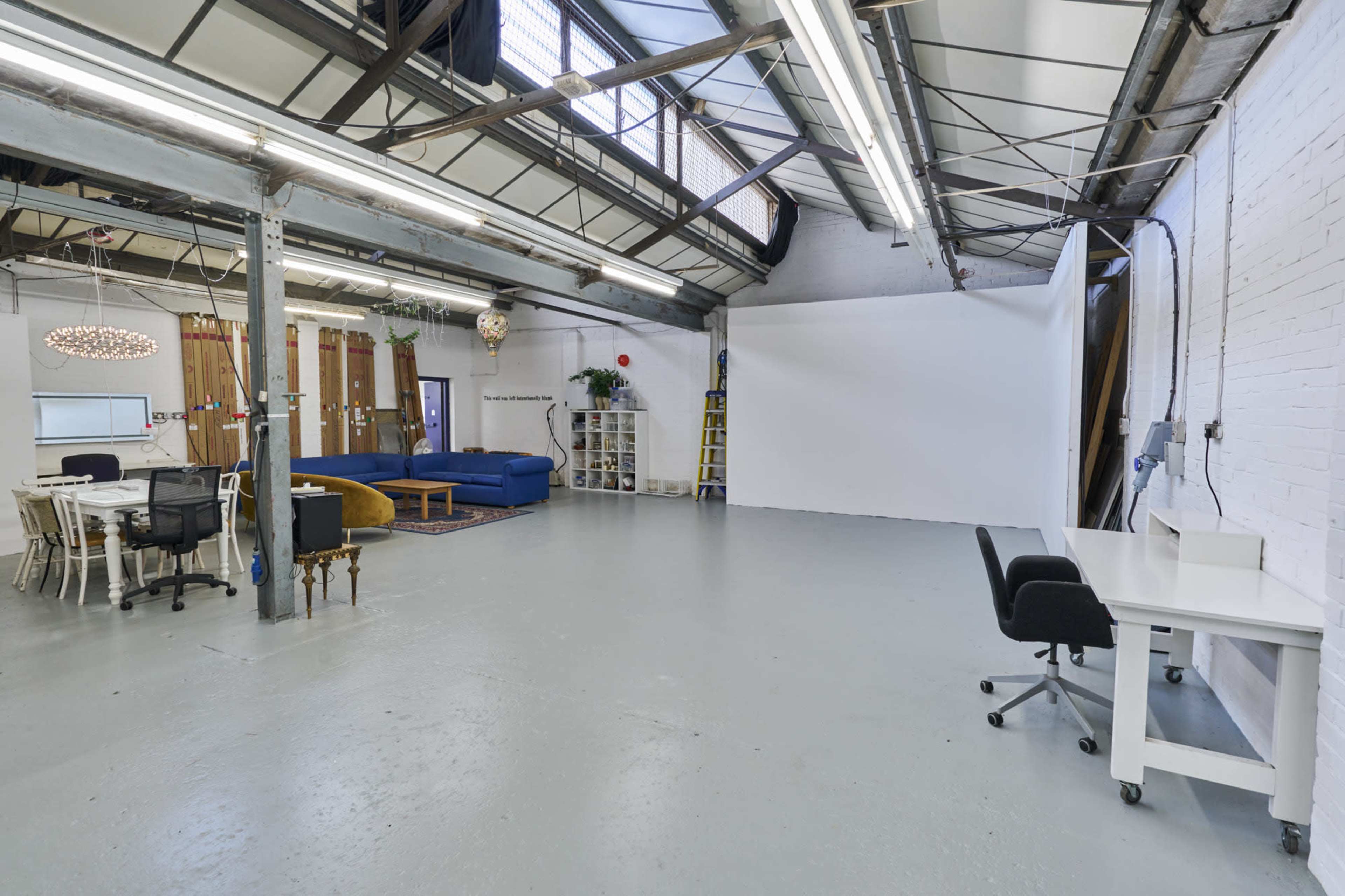 The image shows a spacious, industrial-style room with high ceilings, a large white backdrop, and various furniture pieces including a dining table, chairs, and a blue couch.