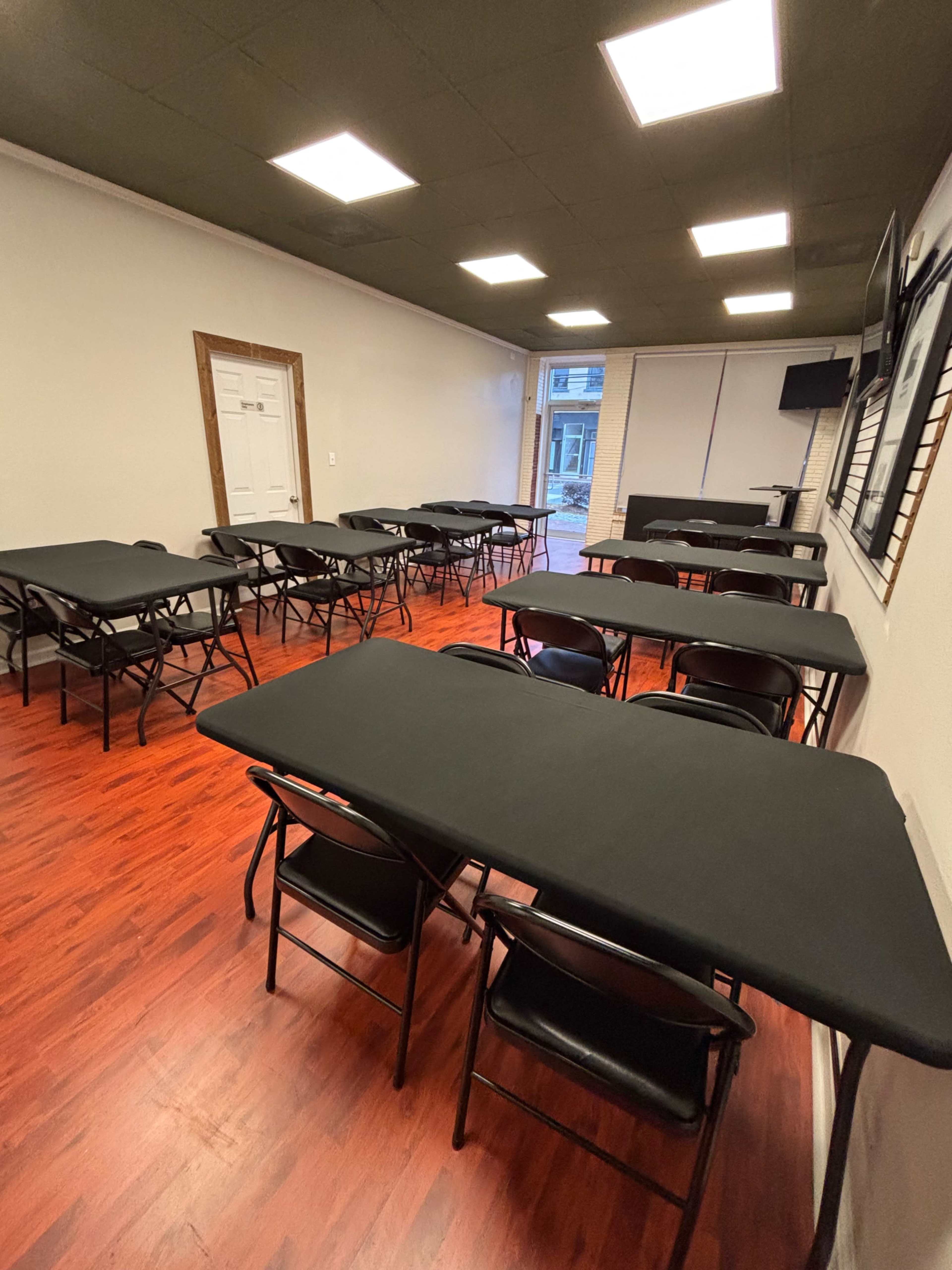 The image shows a room with multiple black folding tables arranged in rows on a wooden floor, with overhead lighting illuminating the space.