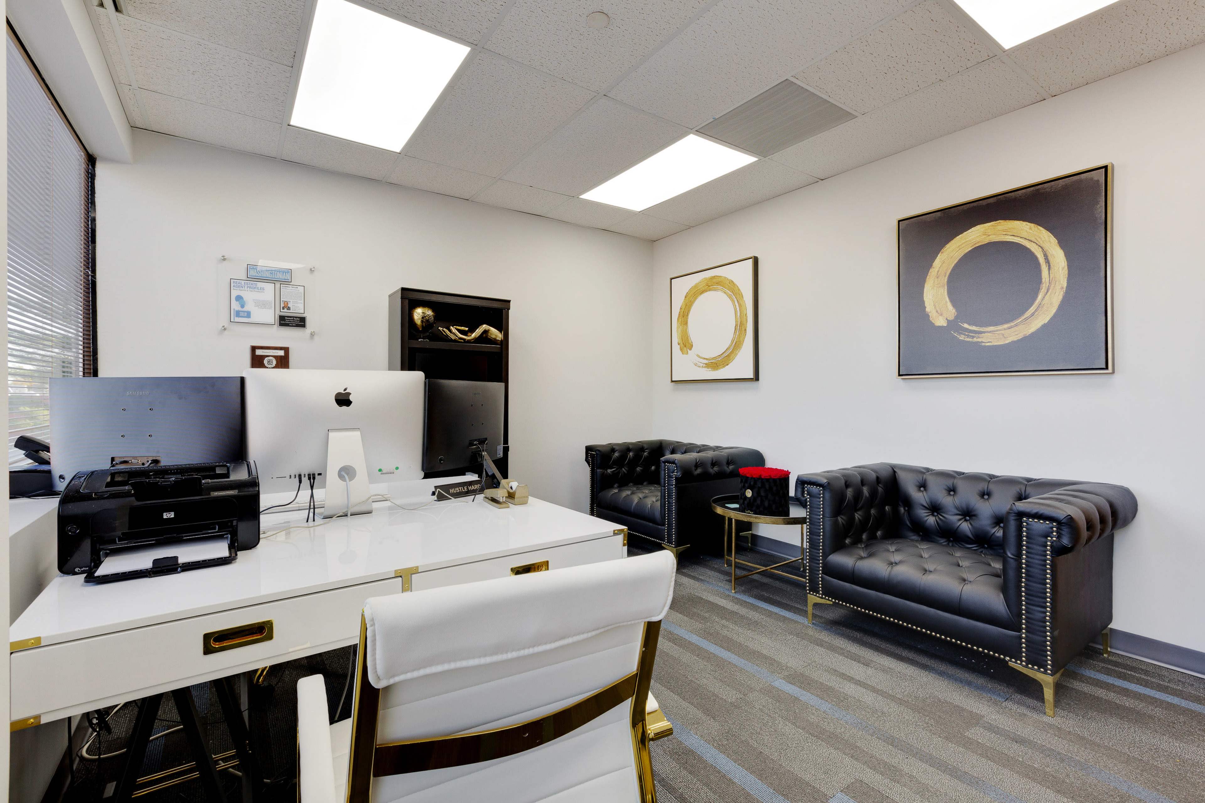 A modern office space featuring a white desk, two black tufted couches, and abstract gold circle art on the walls.