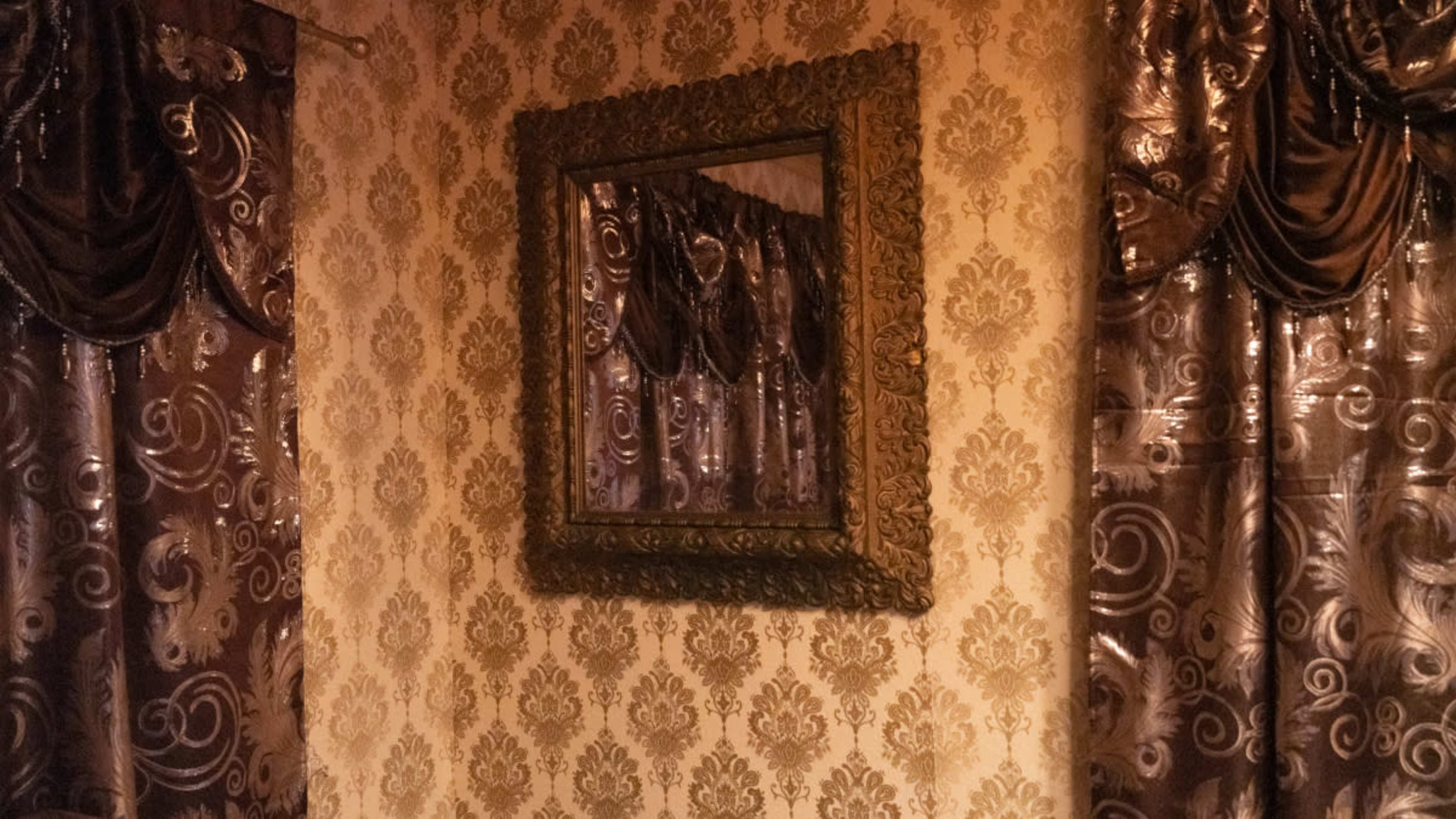 A decorative mirror is hung on a patterned wall surrounded by ornate, dark curtains.