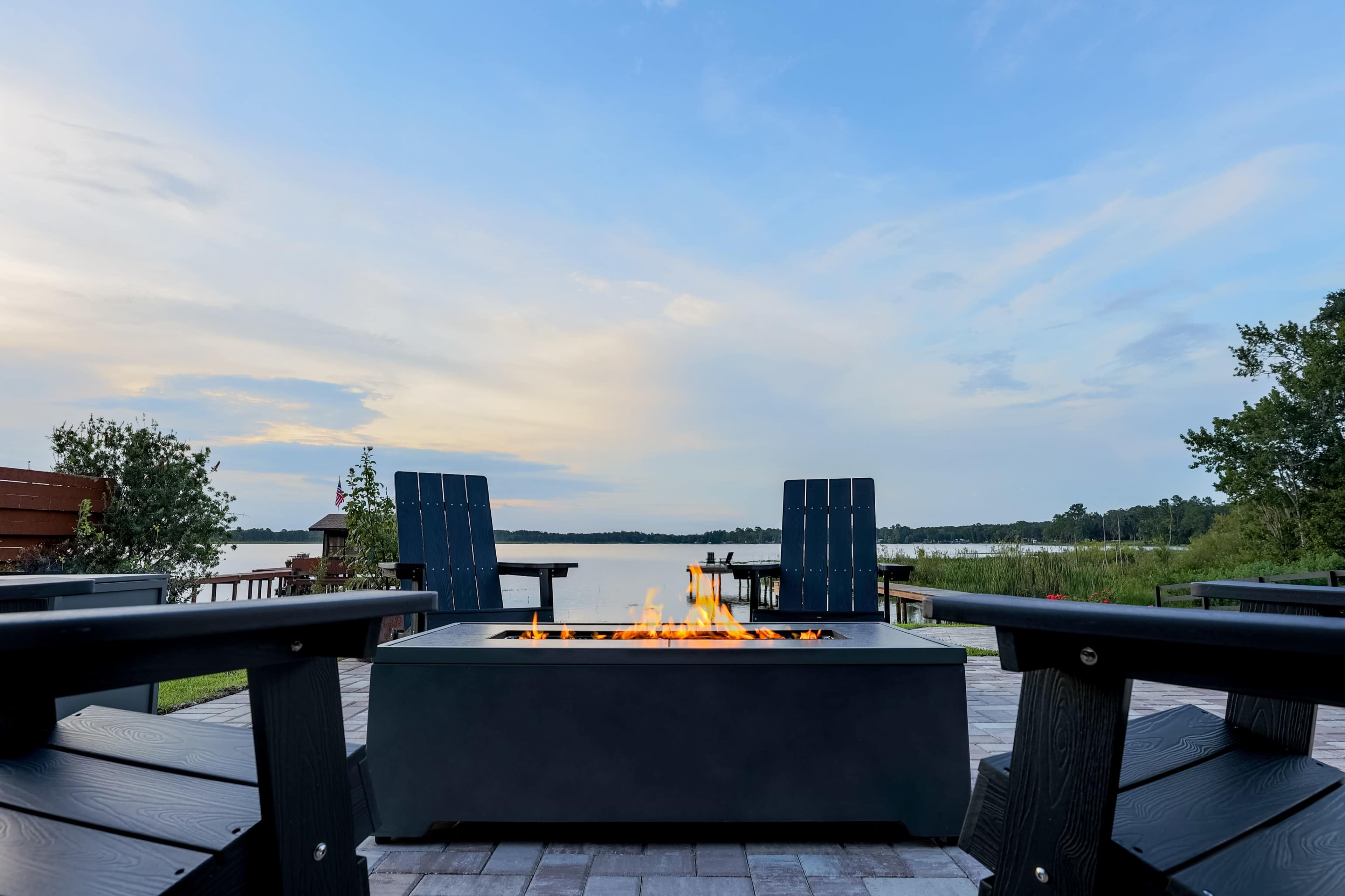 A lakefront patio with an open fire pit surrounded by black lounge chairs, under a blue sky.