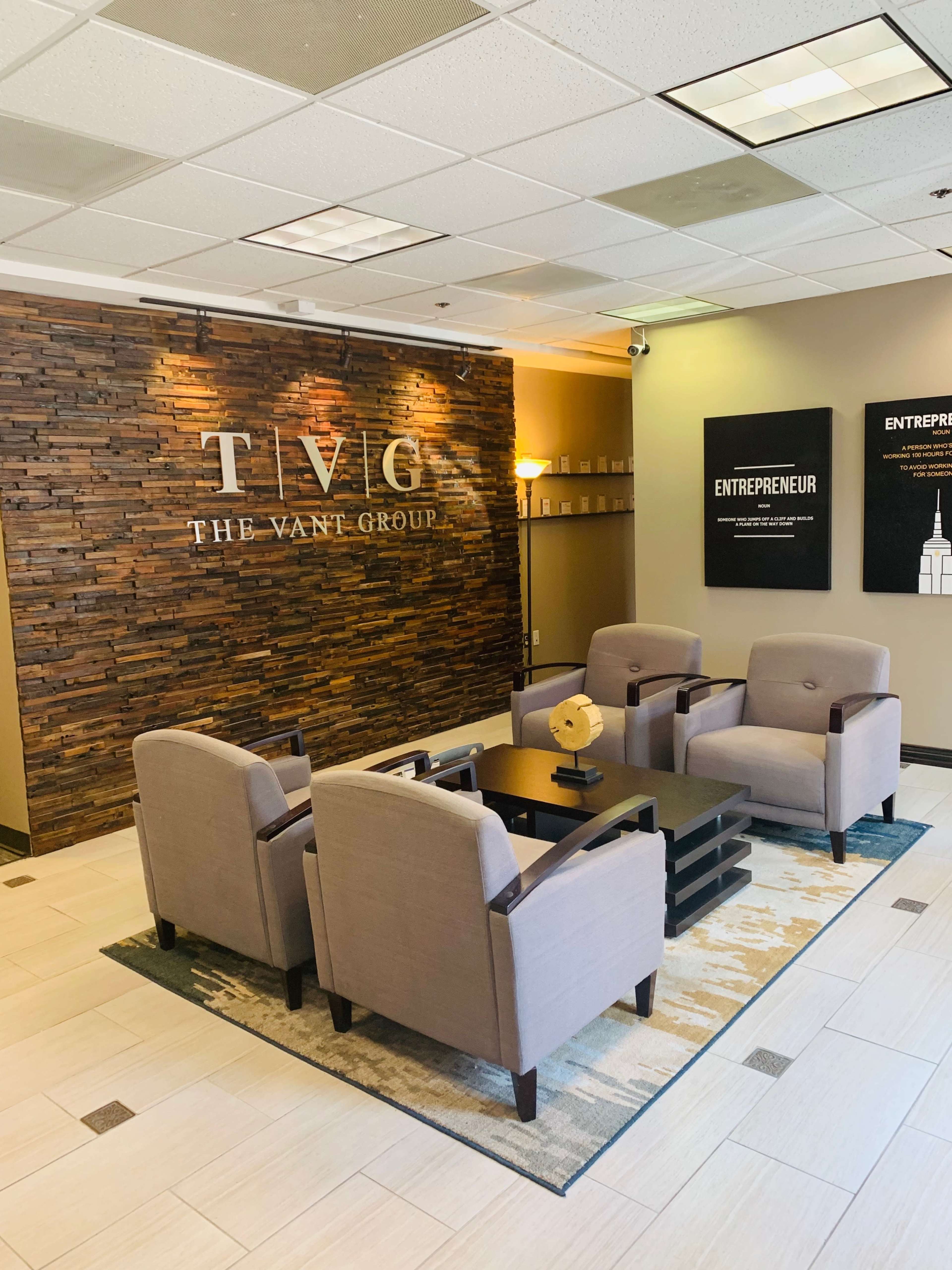 The image shows a modern office waiting area with gray armchairs, a dark coffee table, and a textured wooden wall featuring the name "TVG" and "THE VANT GROUP."