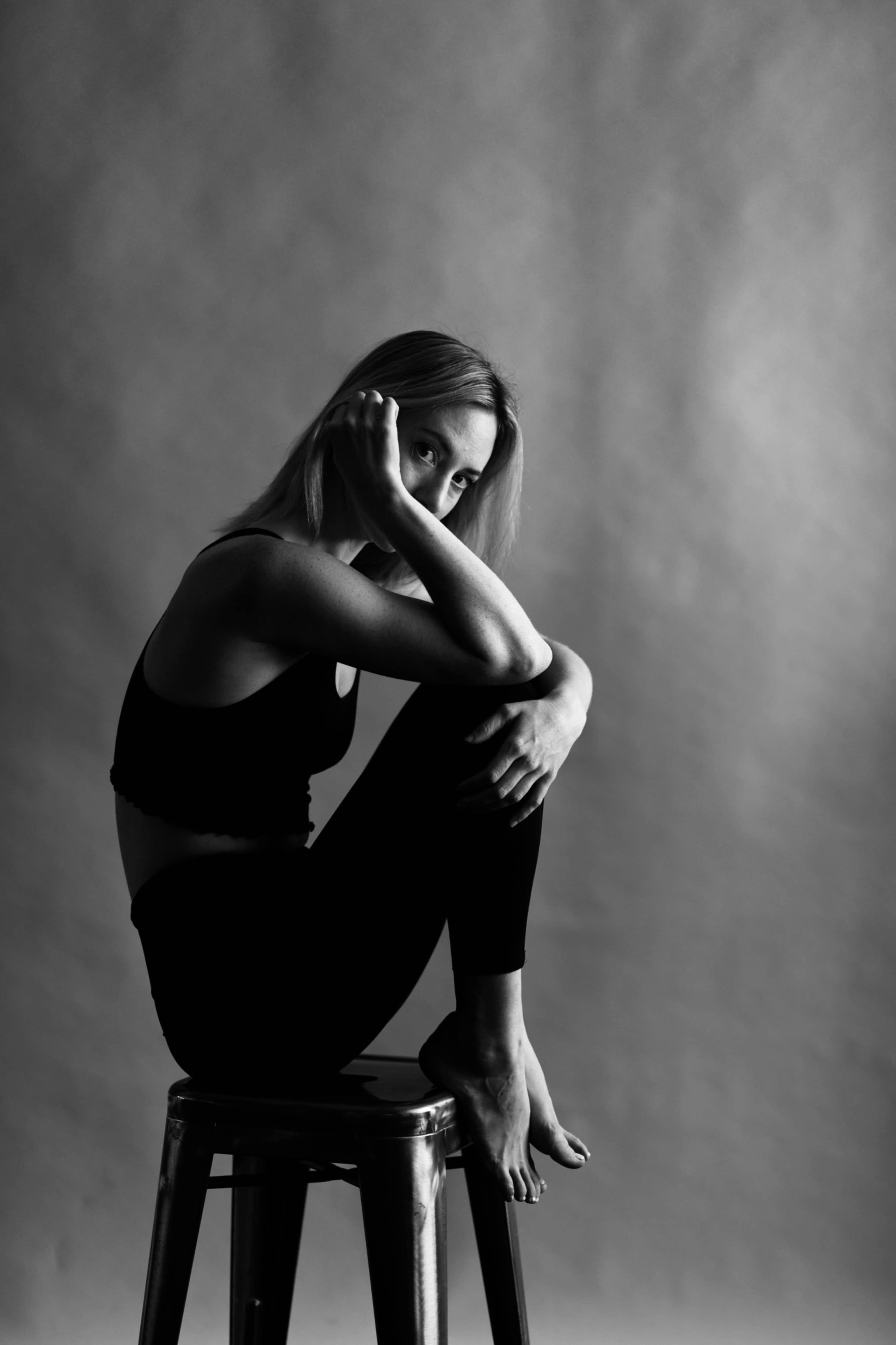 A woman sits on a metal stool with her knees drawn up, resting her chin on her hand against a plain background.
