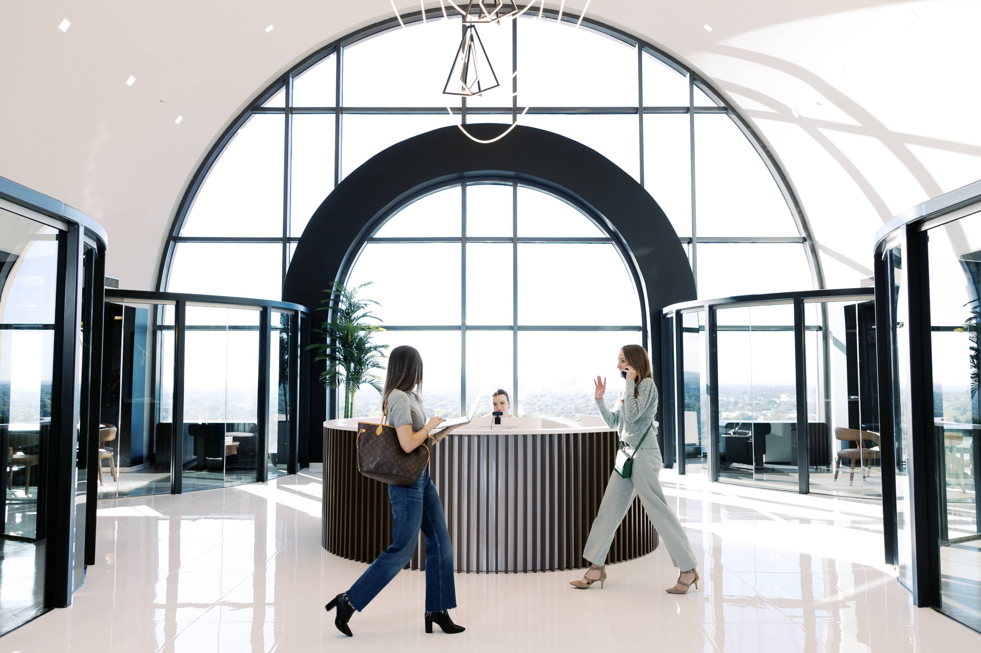Two women walk past a reception desk in a modern lobby featuring large windows and a sleek design.