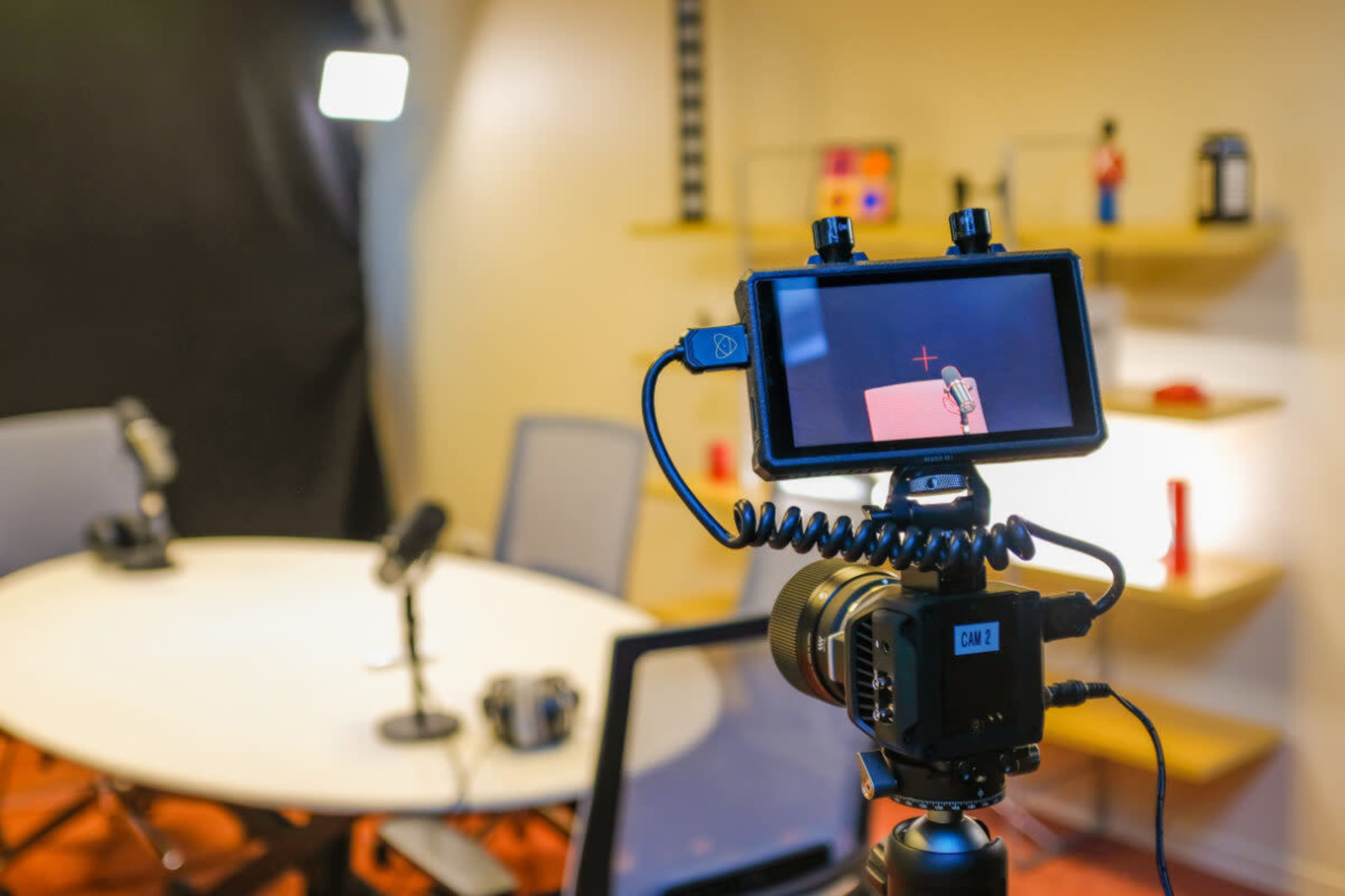 A camera is set up on a tripod, displaying a red setup within a recording studio featuring microphones and a round table.