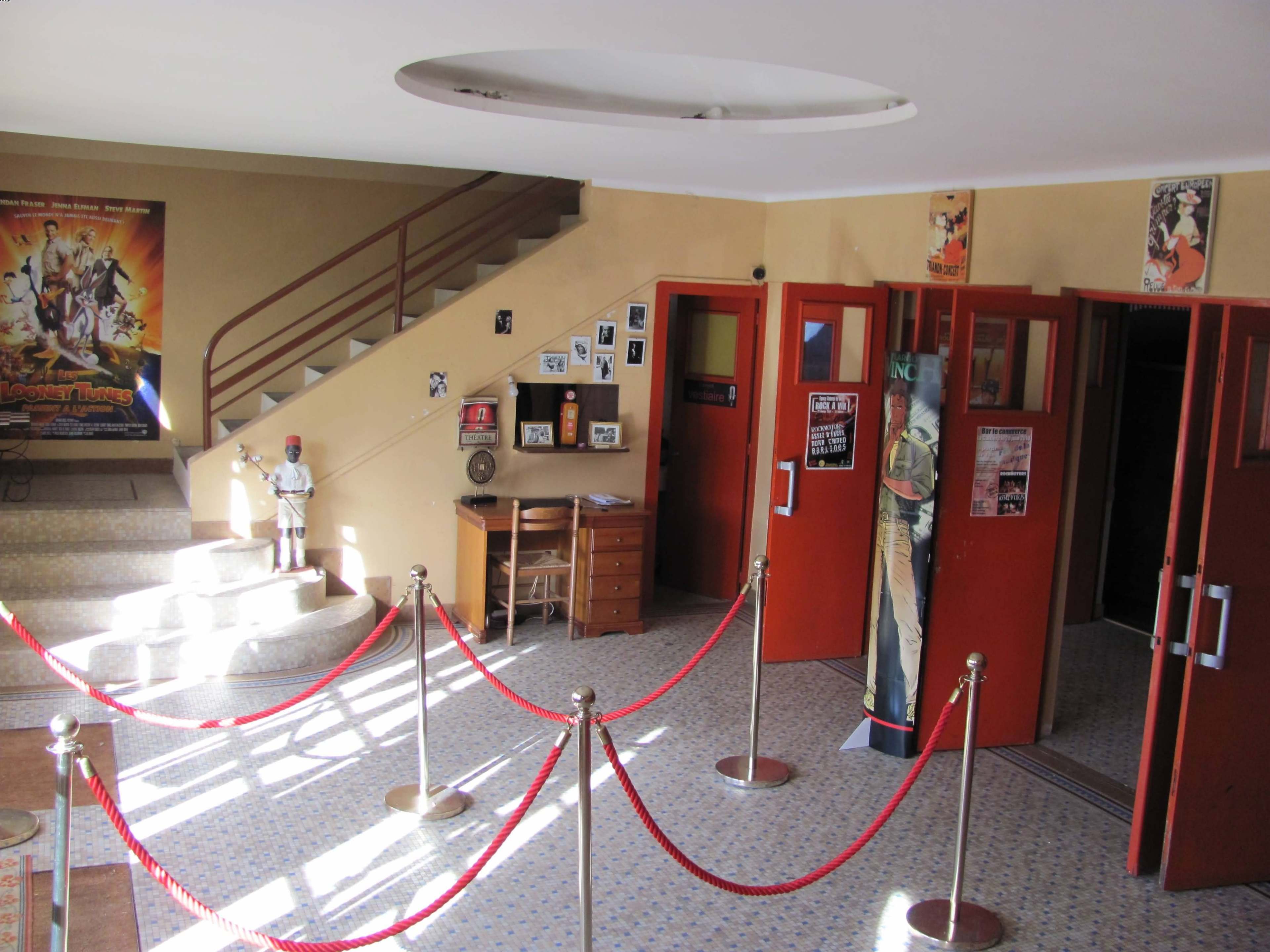 An interior view of a vintage movie theater lobby with a staircase, red velvet ropes, and posters on the walls.