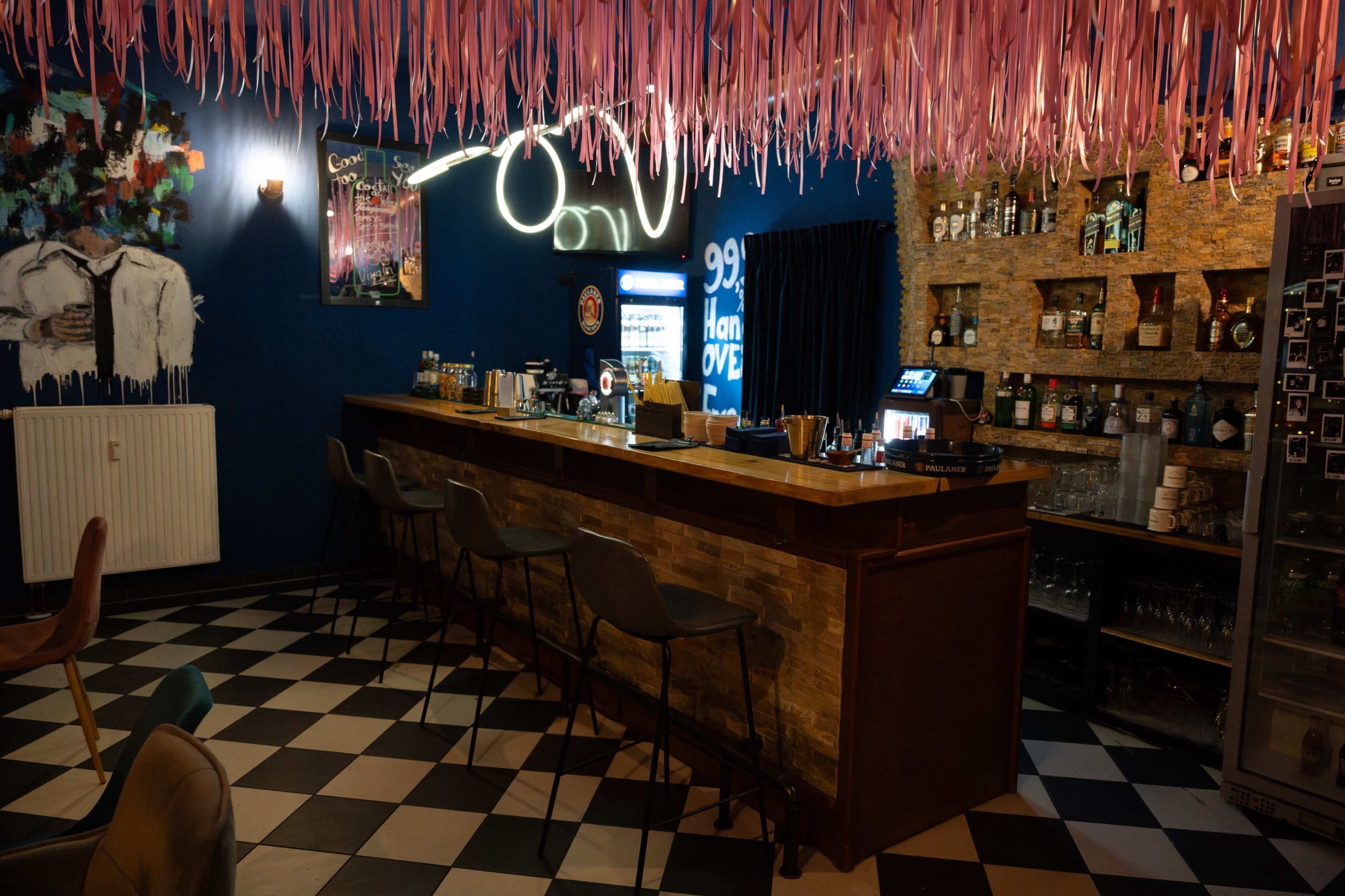 The image shows a bar area with a wooden counter, high chairs, and a vibrant pink ceiling decoration, featuring a variety of bottles on a stone wall behind the bar.