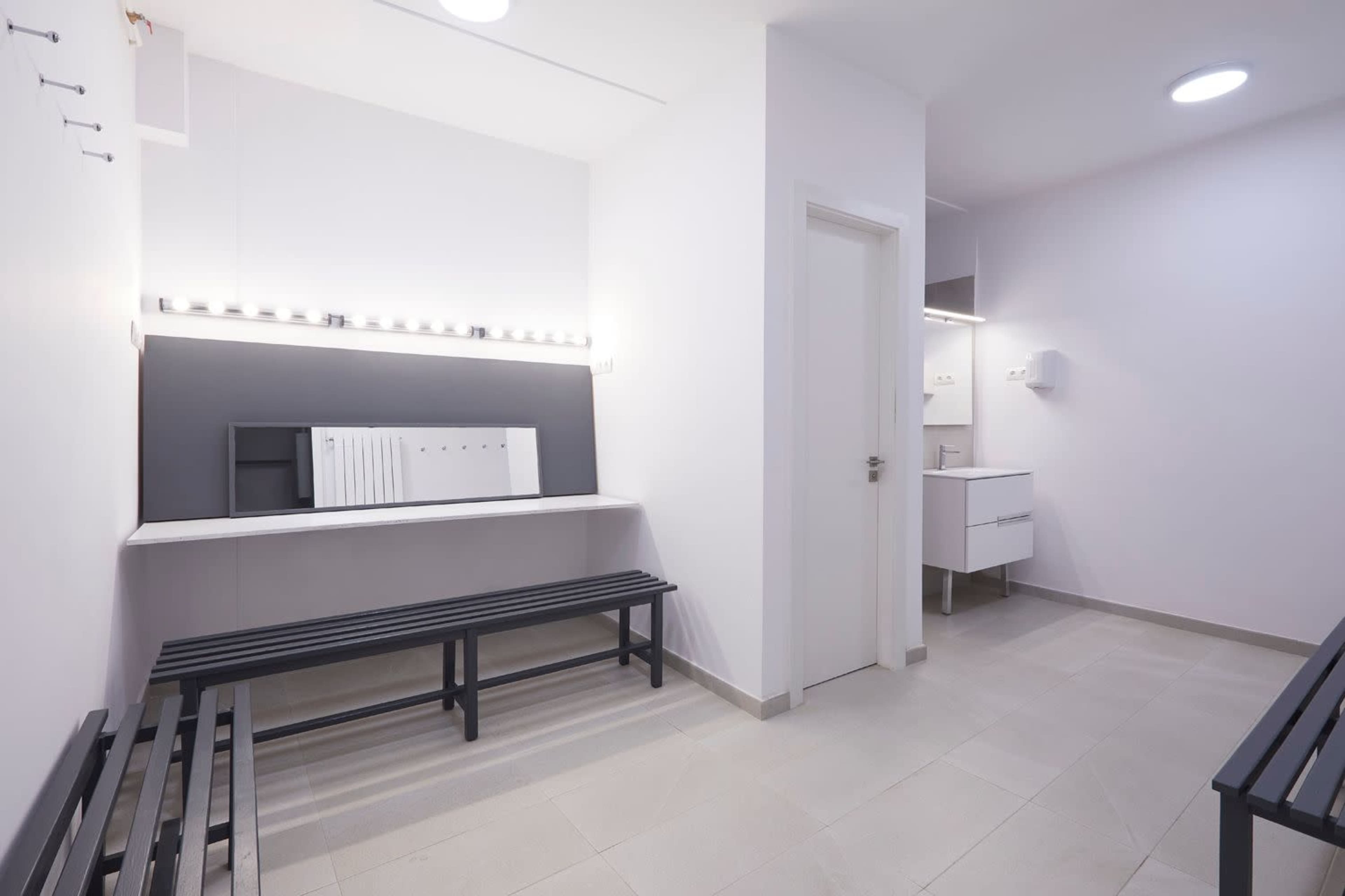 A minimalist waiting area with two benches, a lighted mirror, and a small sink area in a clean, modern design.