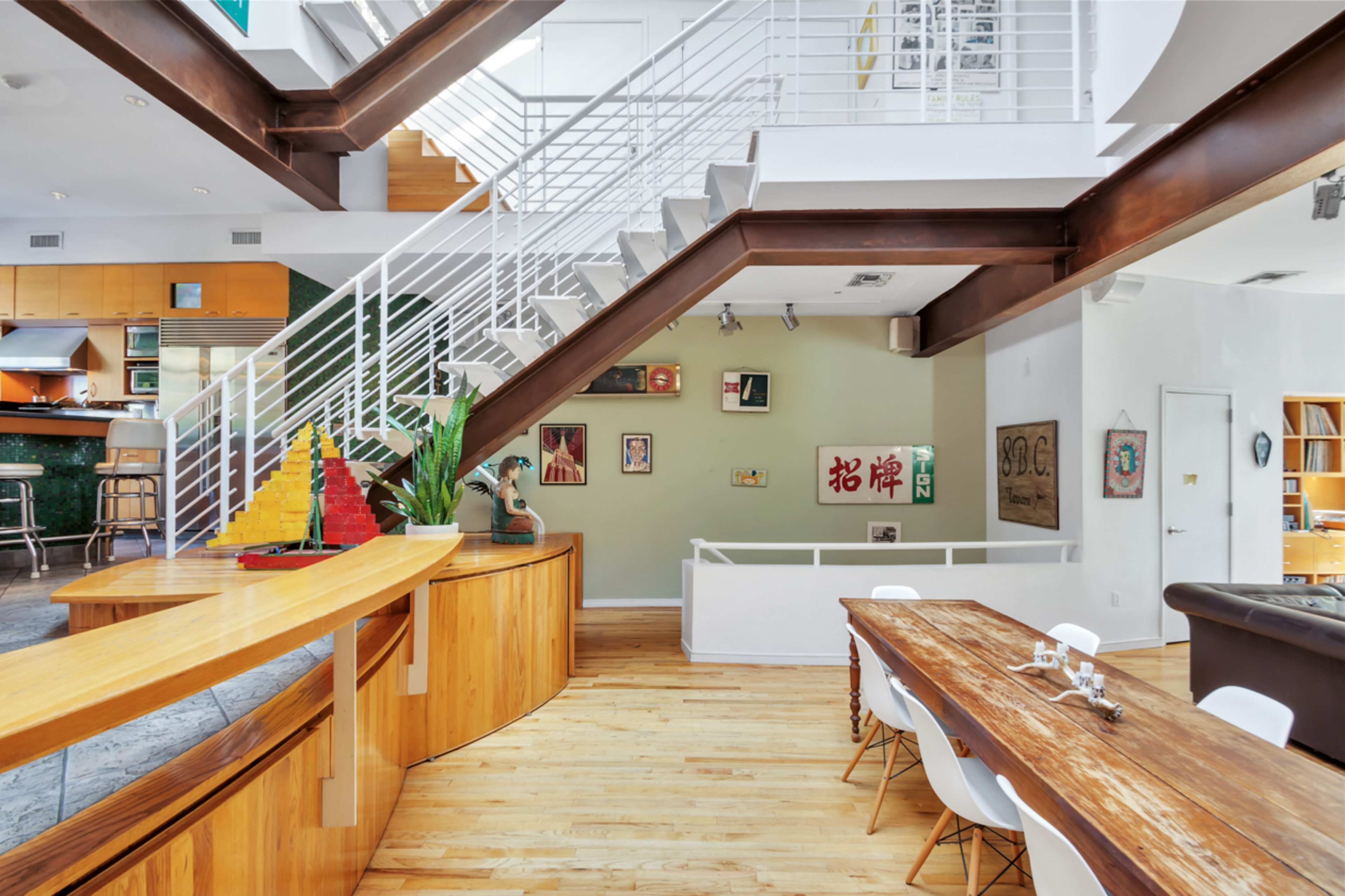 A spacious, open-concept living area with a staircase, wooden dining table, and various artworks displayed on the walls.