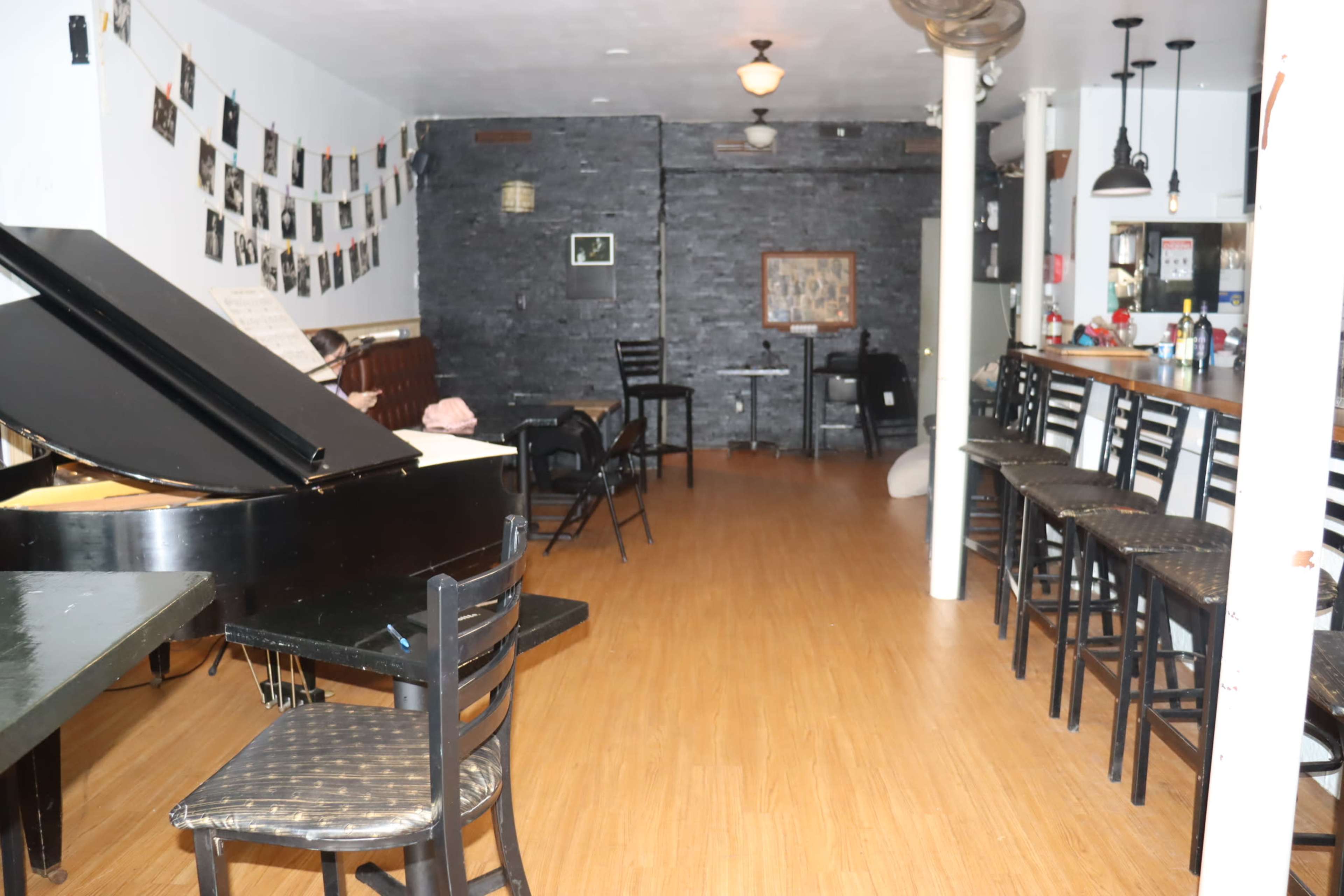 The interior of a music venue features a grand piano in the foreground, rows of black chairs, and a bar against the back wall.