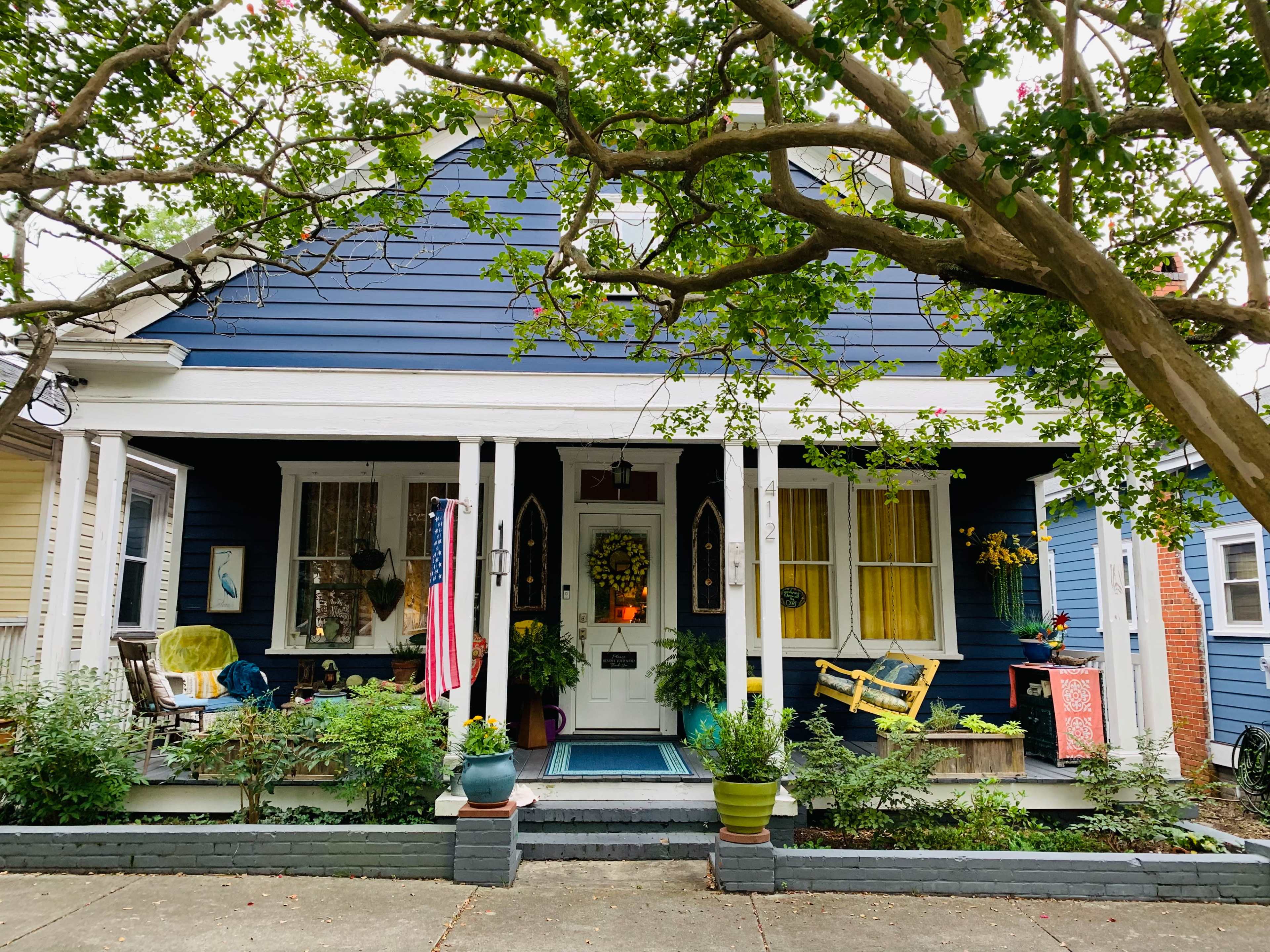 A blue house with white columns and porch, adorned with plants and an American flag, sits beneath a large tree.