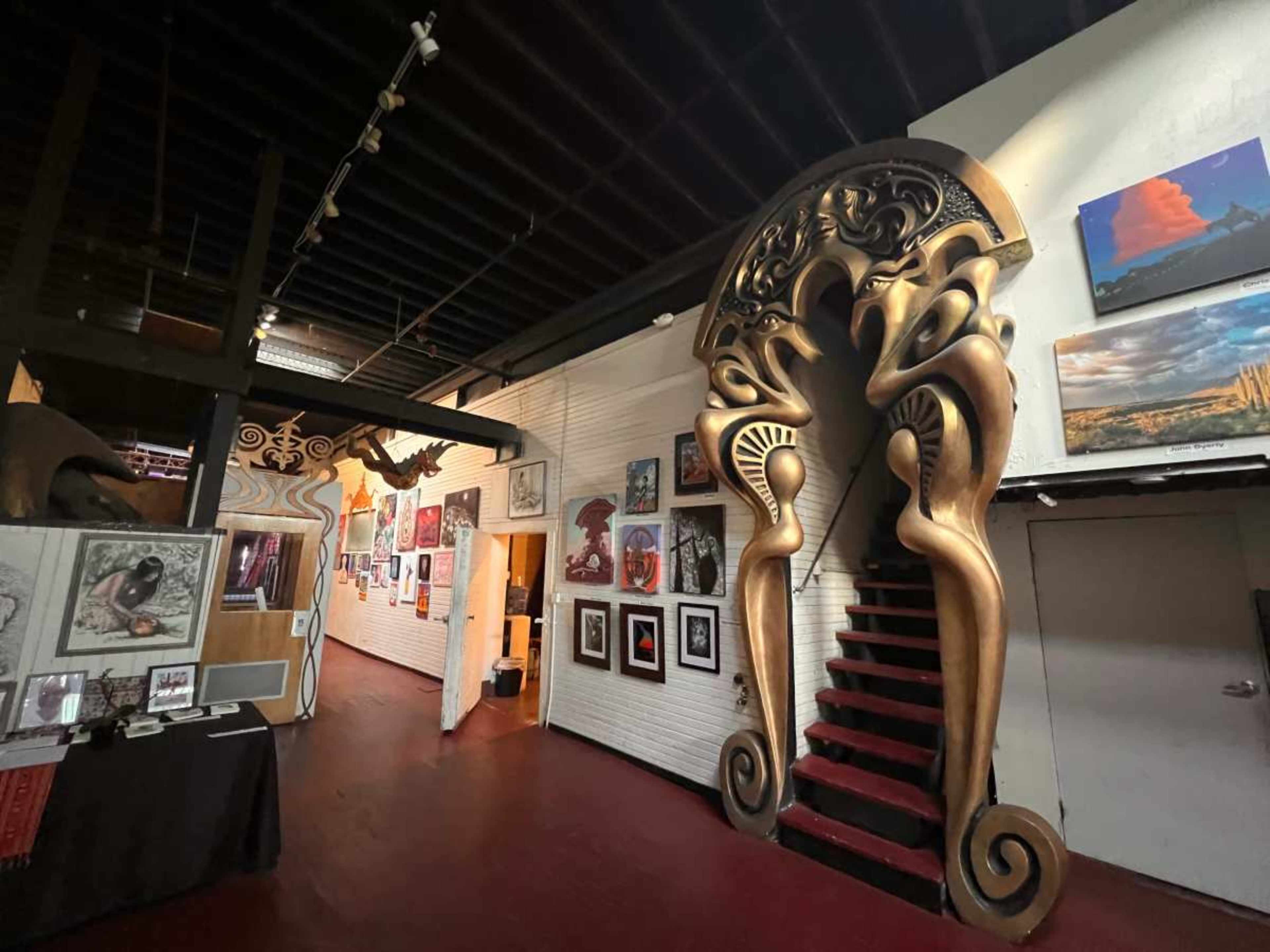 The image shows an art gallery hallway with intricately designed staircases and various artworks displayed on the walls.