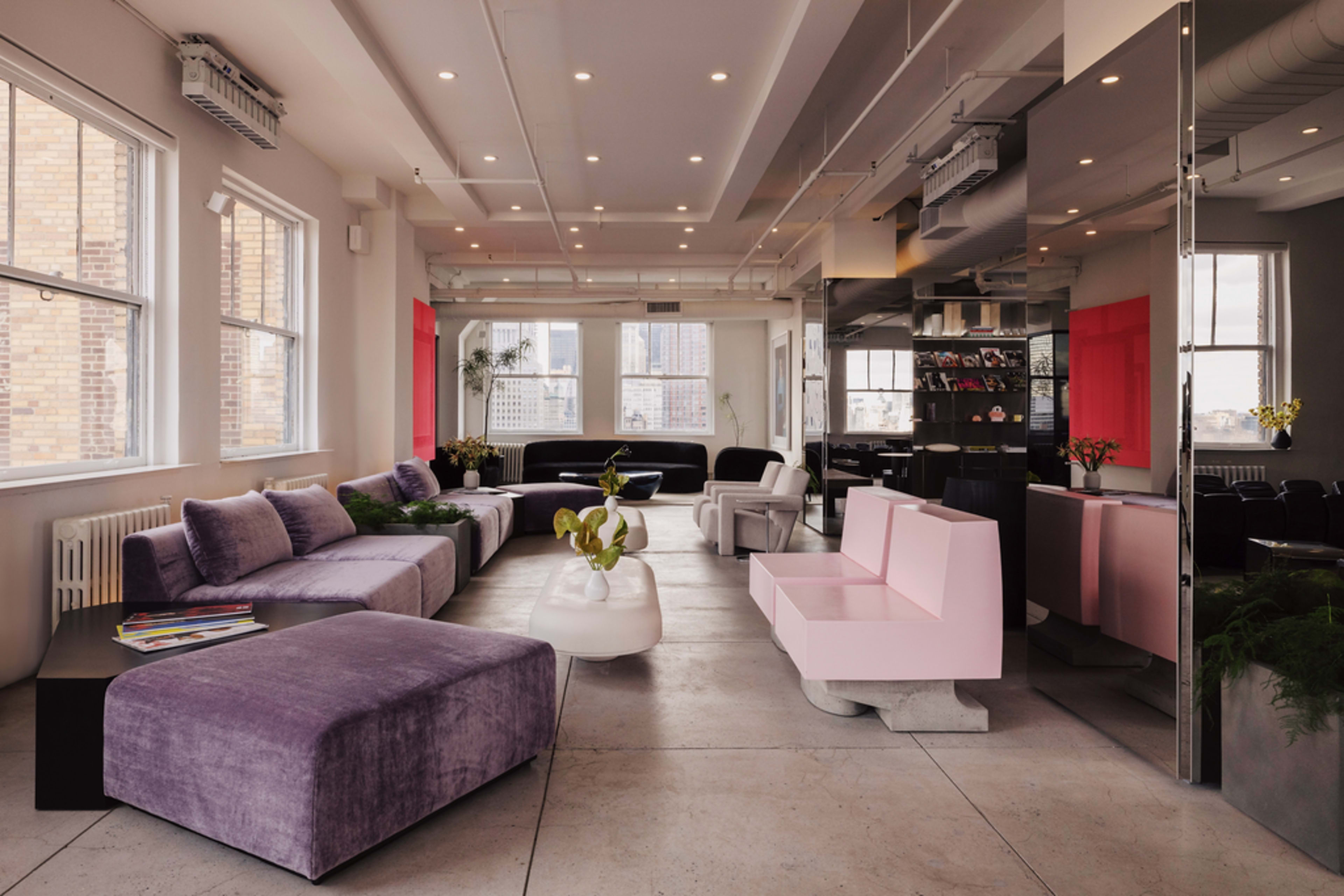 The image shows a modern, stylish lounge area featuring a variety of seating options, including purple and pink sofas, large windows, and minimalist decor.