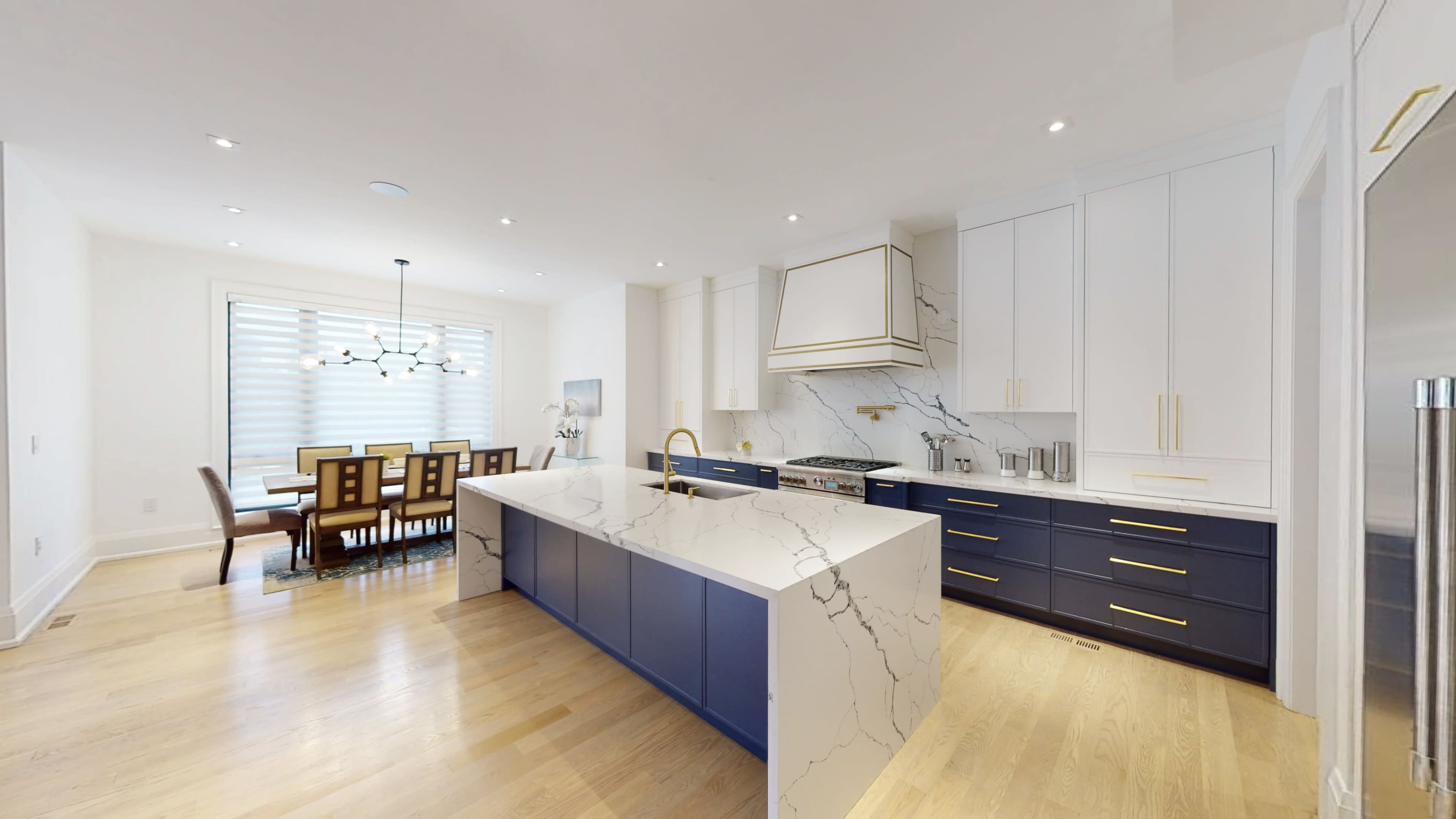 A modern kitchen features a large island with a white and blue marble countertop, surrounded by white cabinetry and gold accents, with a dining area visible in the background.