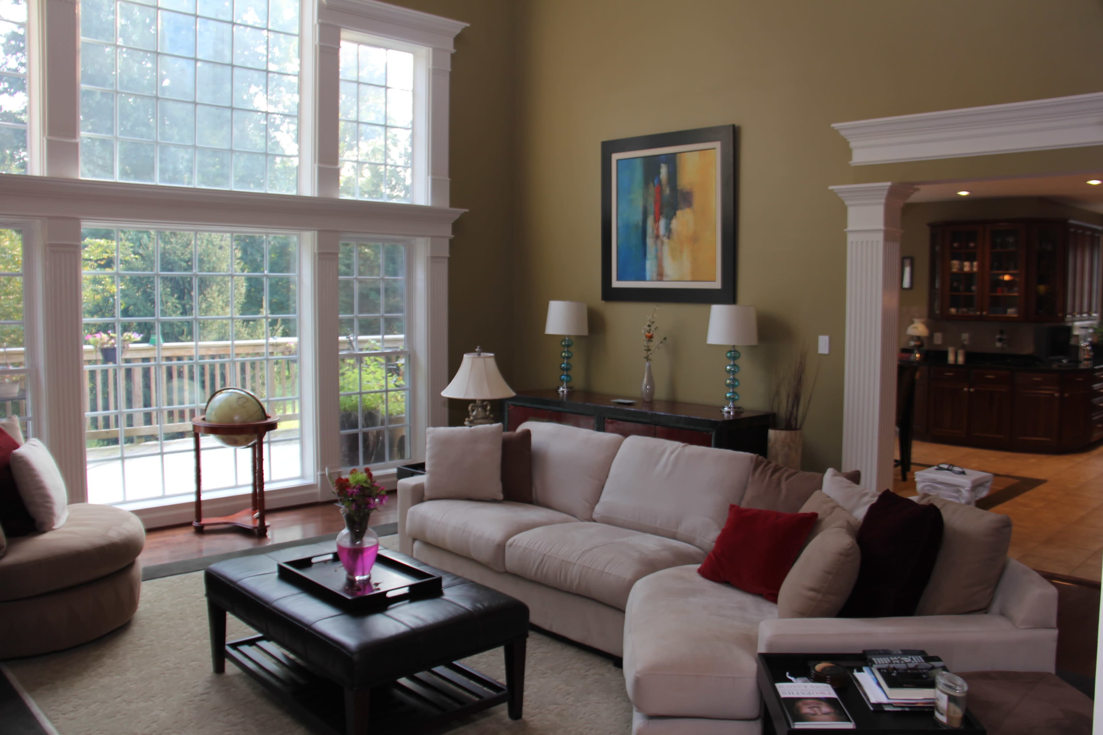 A spacious living room with large windows, a cream-colored sectional sofa, and a wooden table in the center, overlooking a deck area outside.