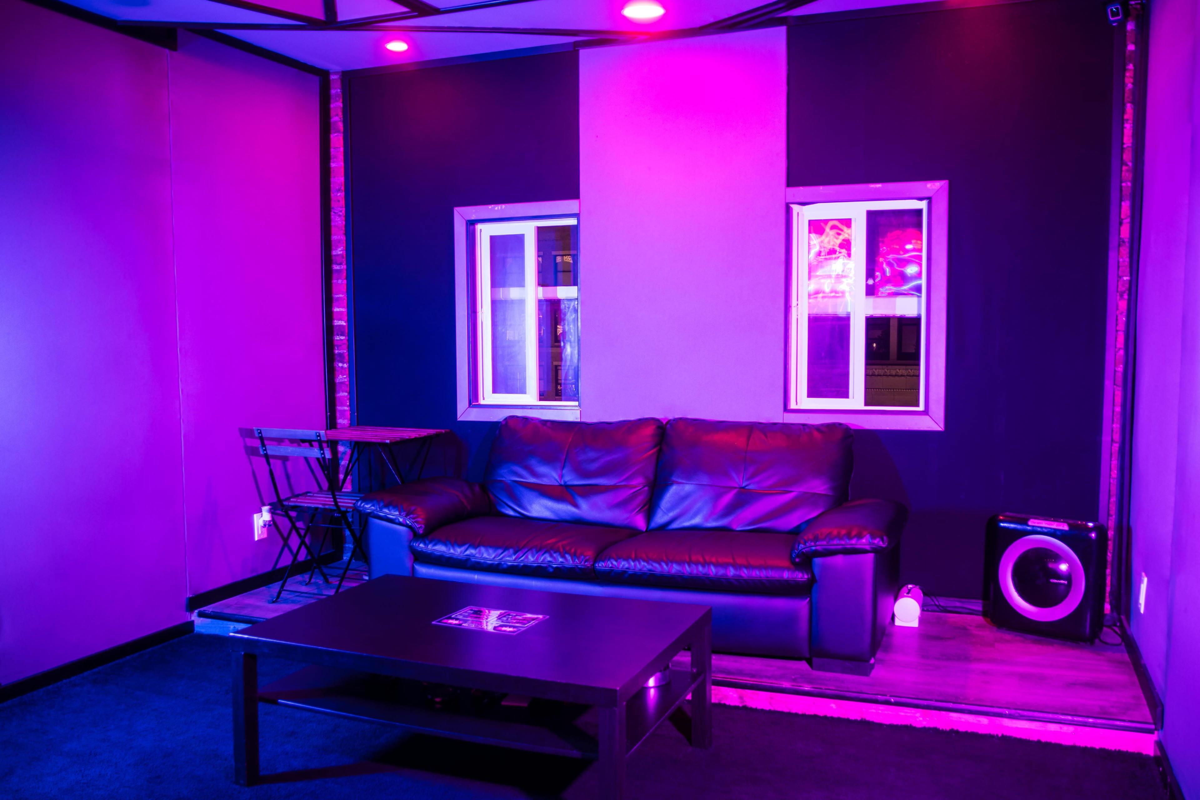 The image shows a dimly lit lounge area with a black leather couch, a small table, and two windows, all against a backdrop of purple and black walls.