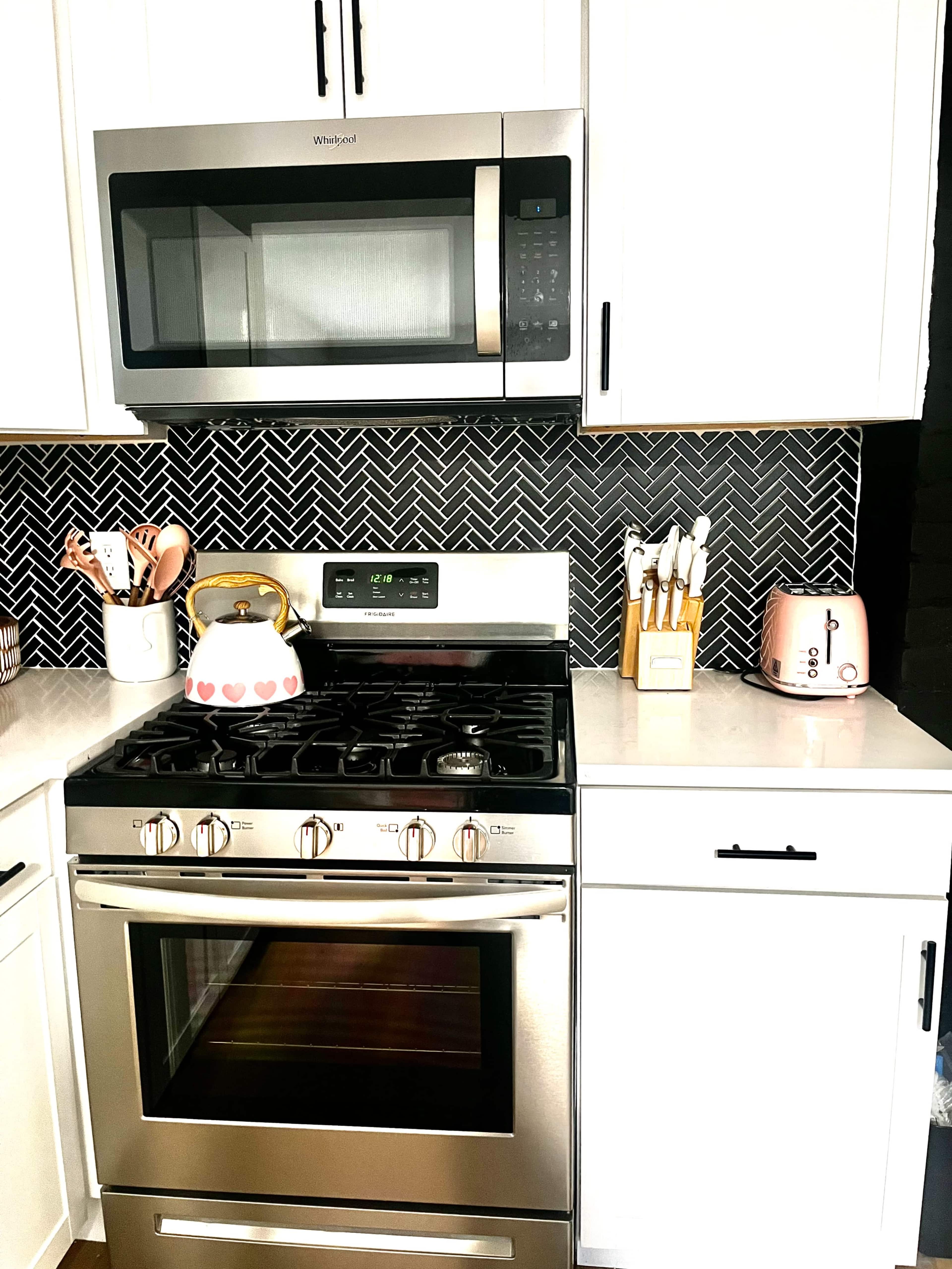 A modern kitchen featuring stainless steel appliances, including a gas stove and microwave, against a patterned backsplash with white cabinets and a pink toaster.
