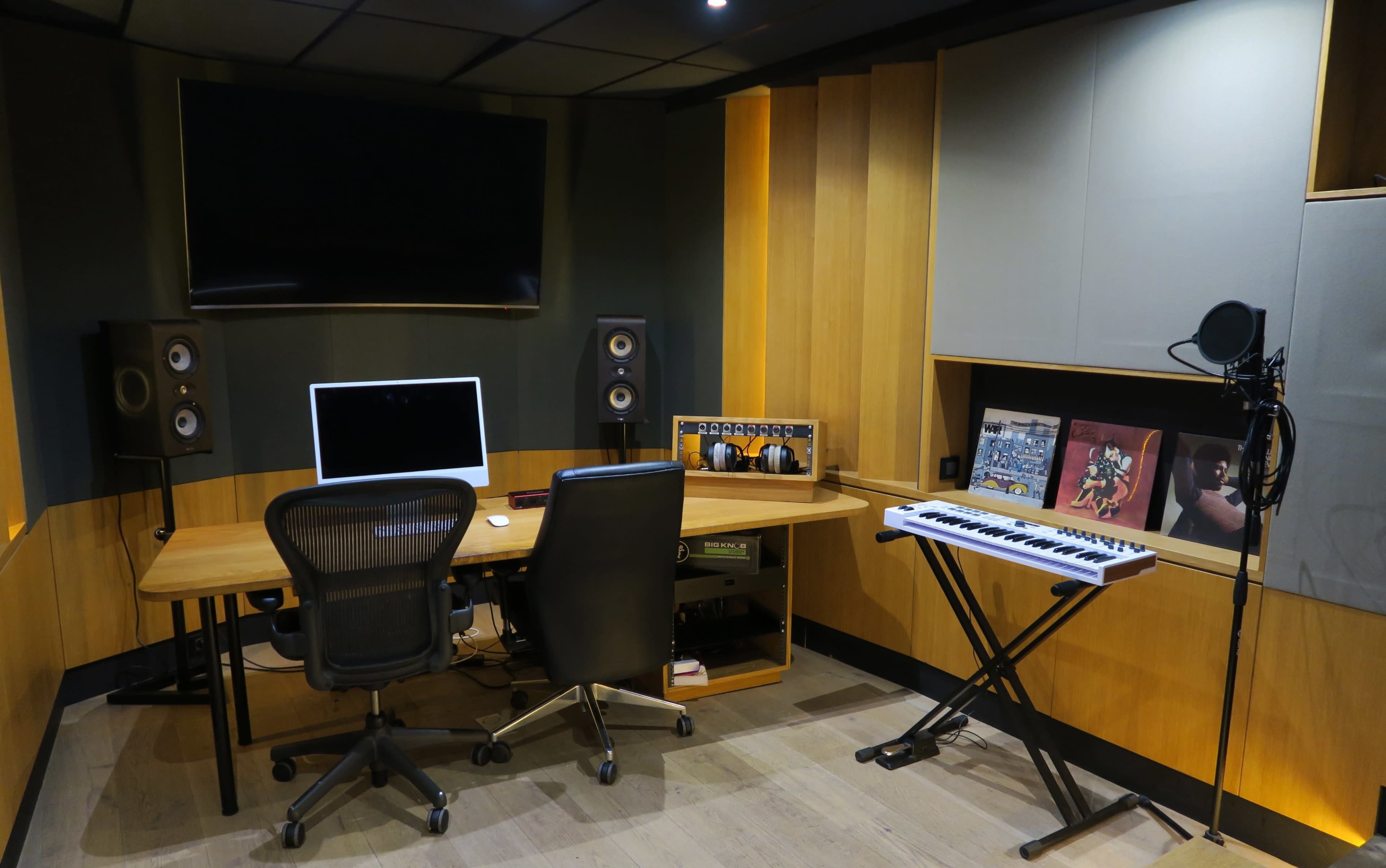 The image shows a music production studio featuring a desk with two chairs, an Apple computer, speakers, and a keyboard, all set against a backdrop of wooden paneling and acoustic treatments.