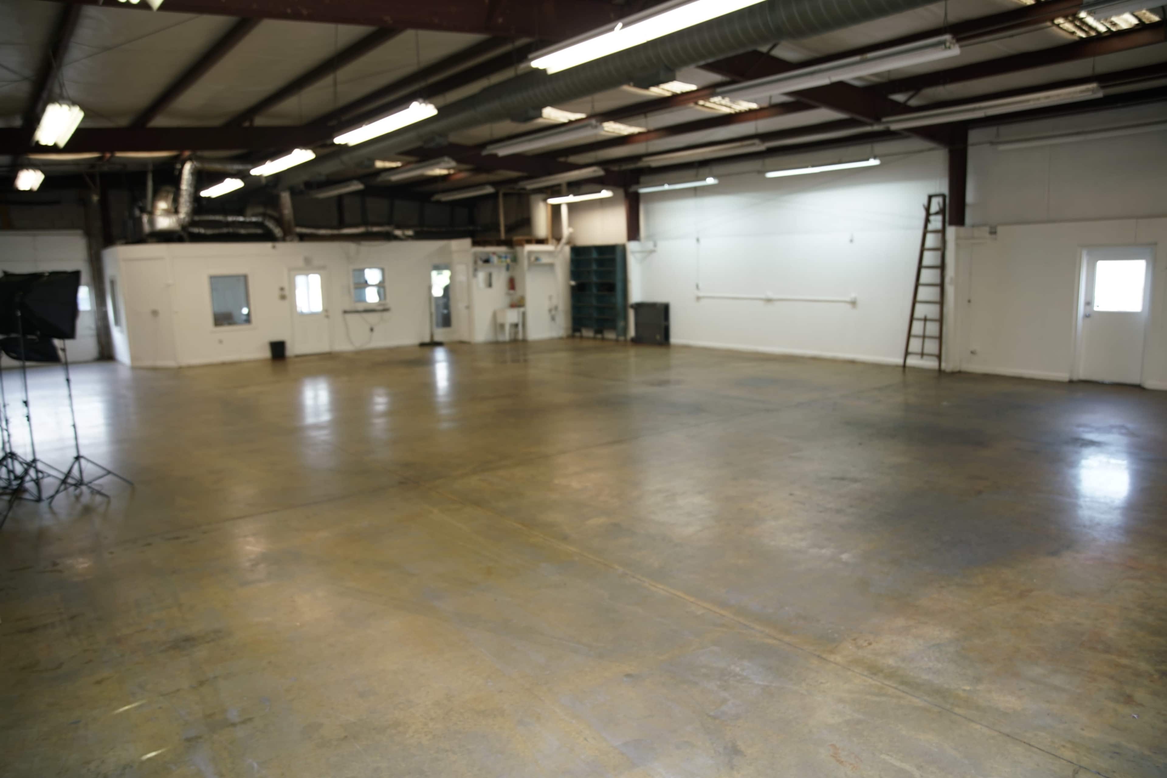 The image shows a spacious industrial room with a concrete floor, several windows, and a ladder against one wall.