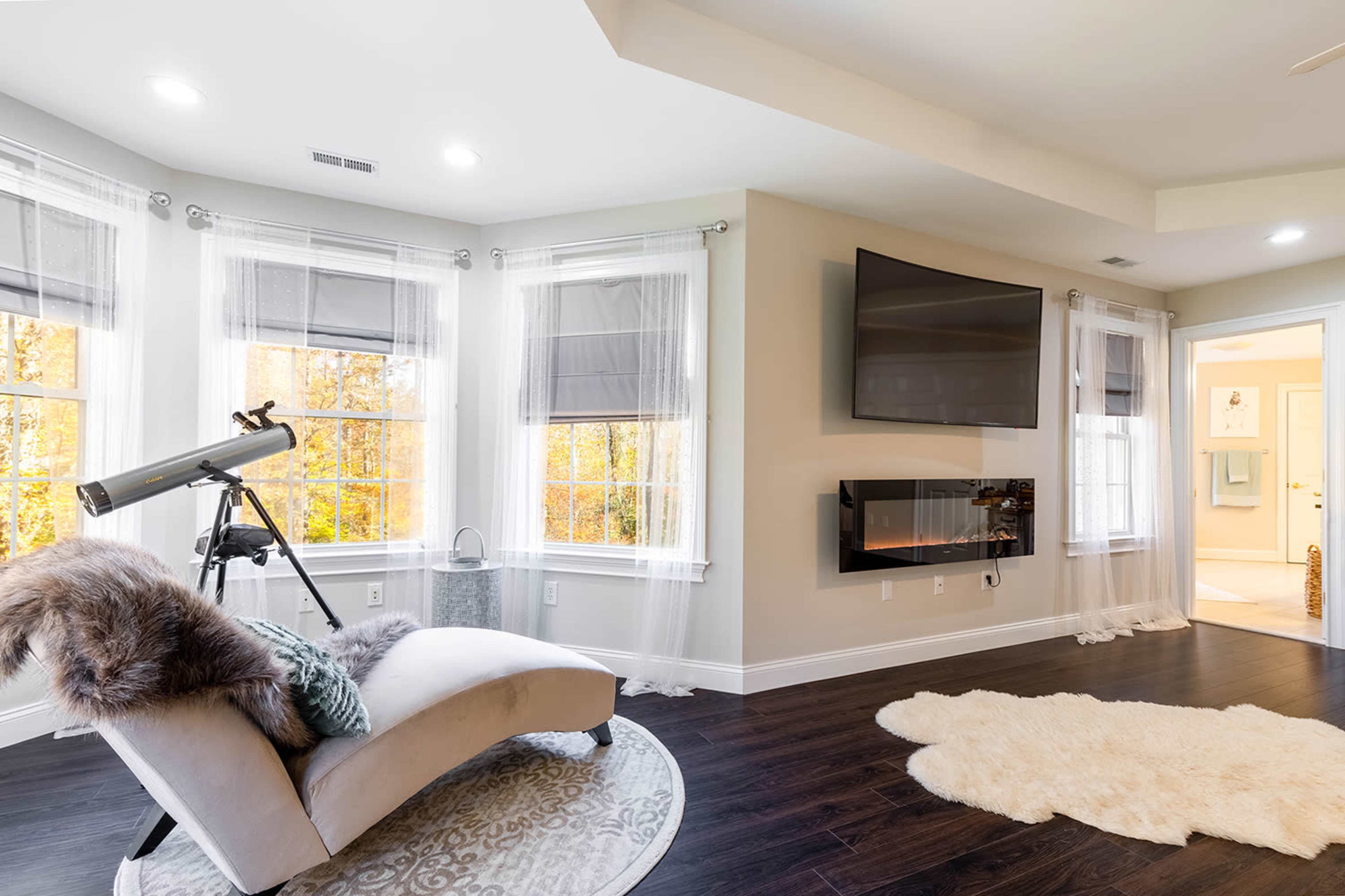 A modern living room features a chaise lounge, a telescope beside it, and large windows draped with sheer curtains, revealing a view of trees outside.
