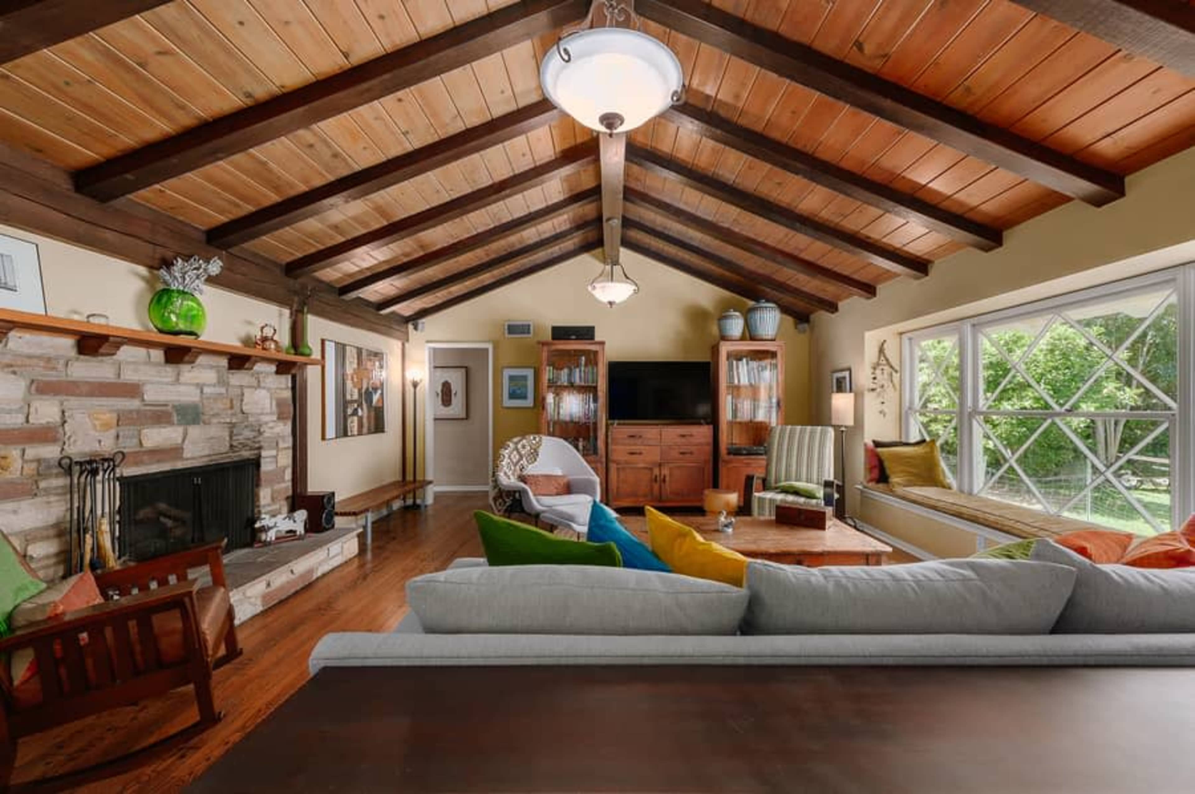 The image shows a spacious living room with wooden beams, a stone fireplace, and large windows letting in natural light.