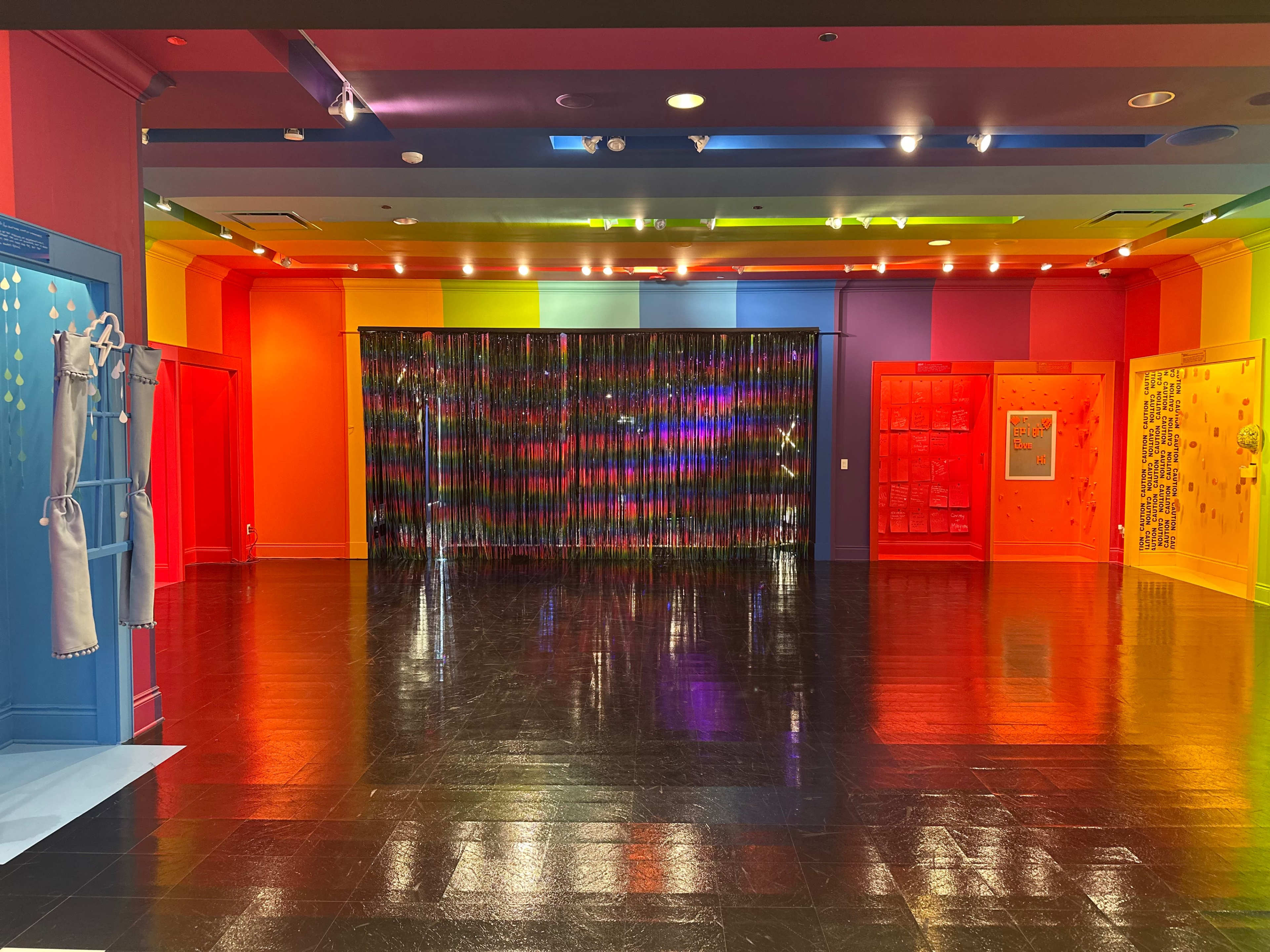 A brightly colored room with walls painted in multiple hues and a shiny black floor, featuring a curtain made of reflective strips at one end.