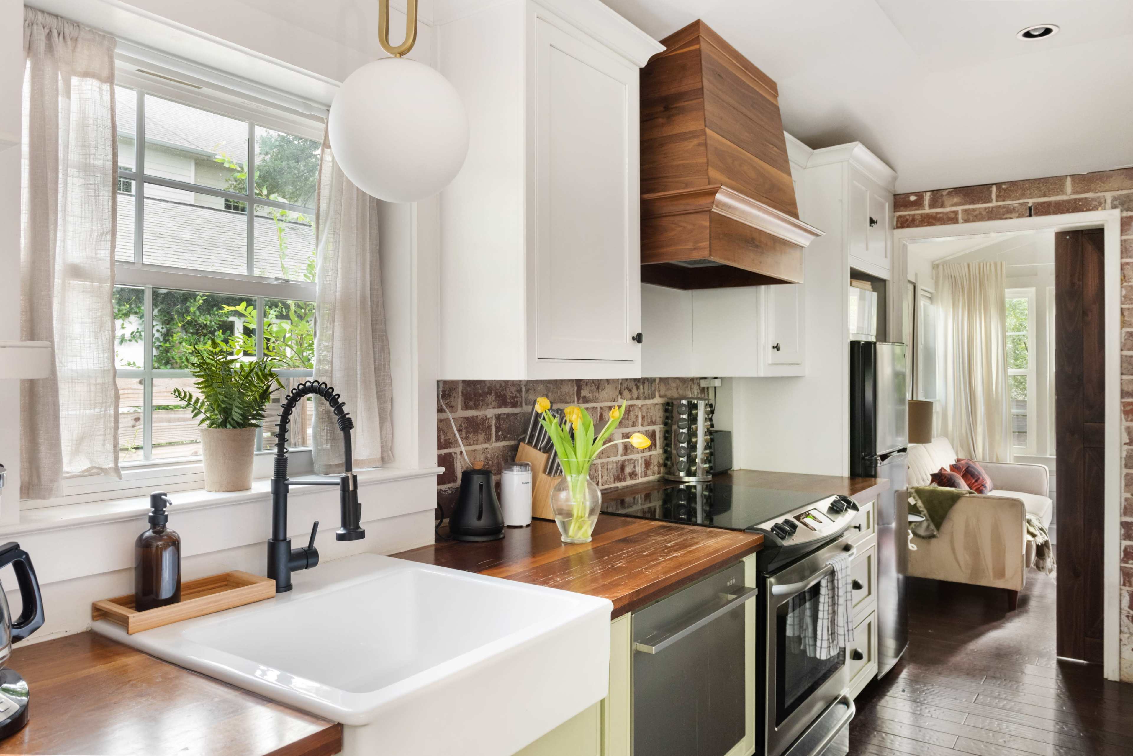 The image shows a modern kitchen with a white farmhouse sink, wooden countertop, and a mix of white and wooden cabinetry, along with a window letting in natural light.