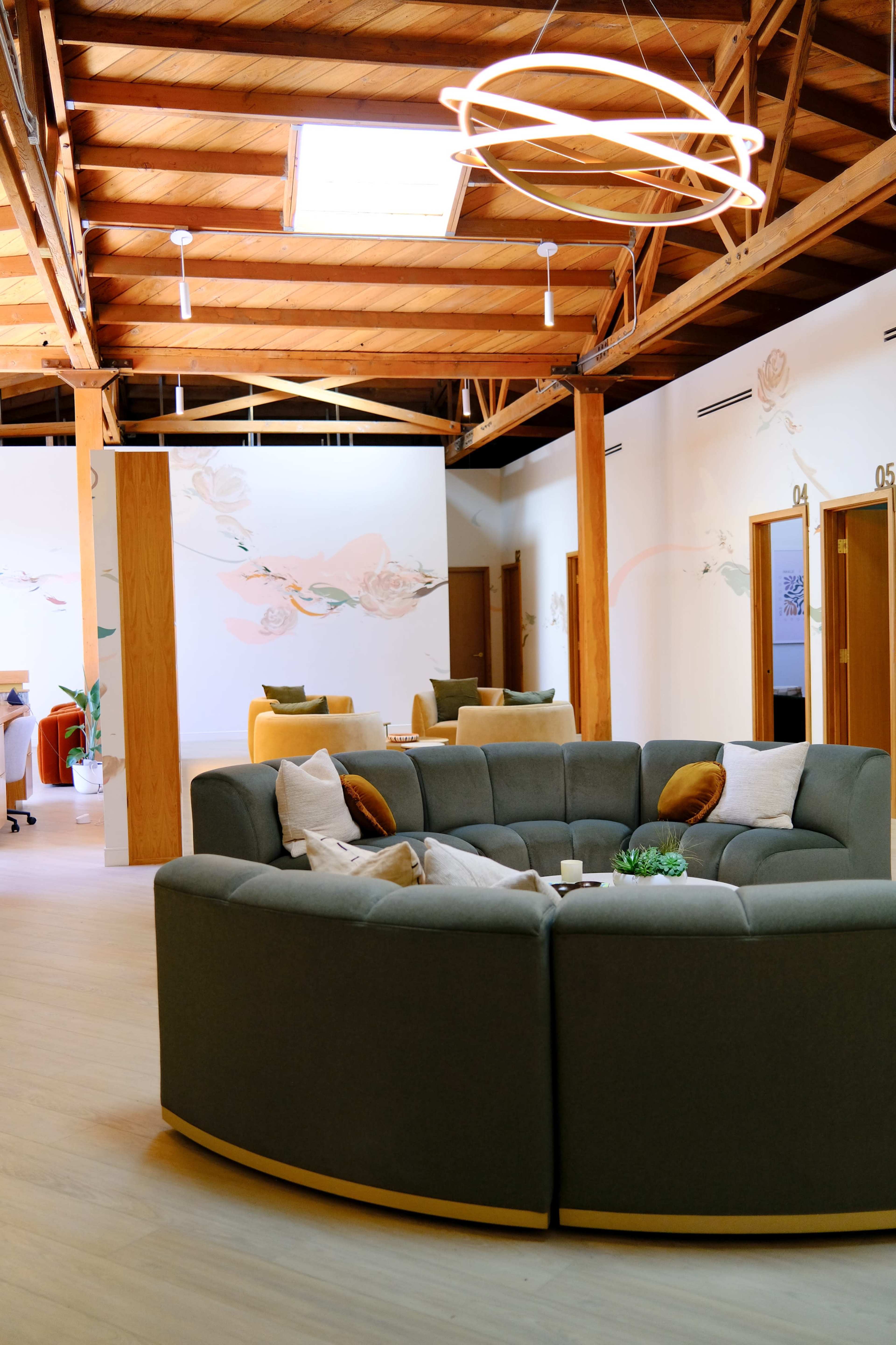 The image shows a modern, circular lounge area with a curved gray sofa and decorative pillows, set in a spacious room featuring wooden beams and light walls.