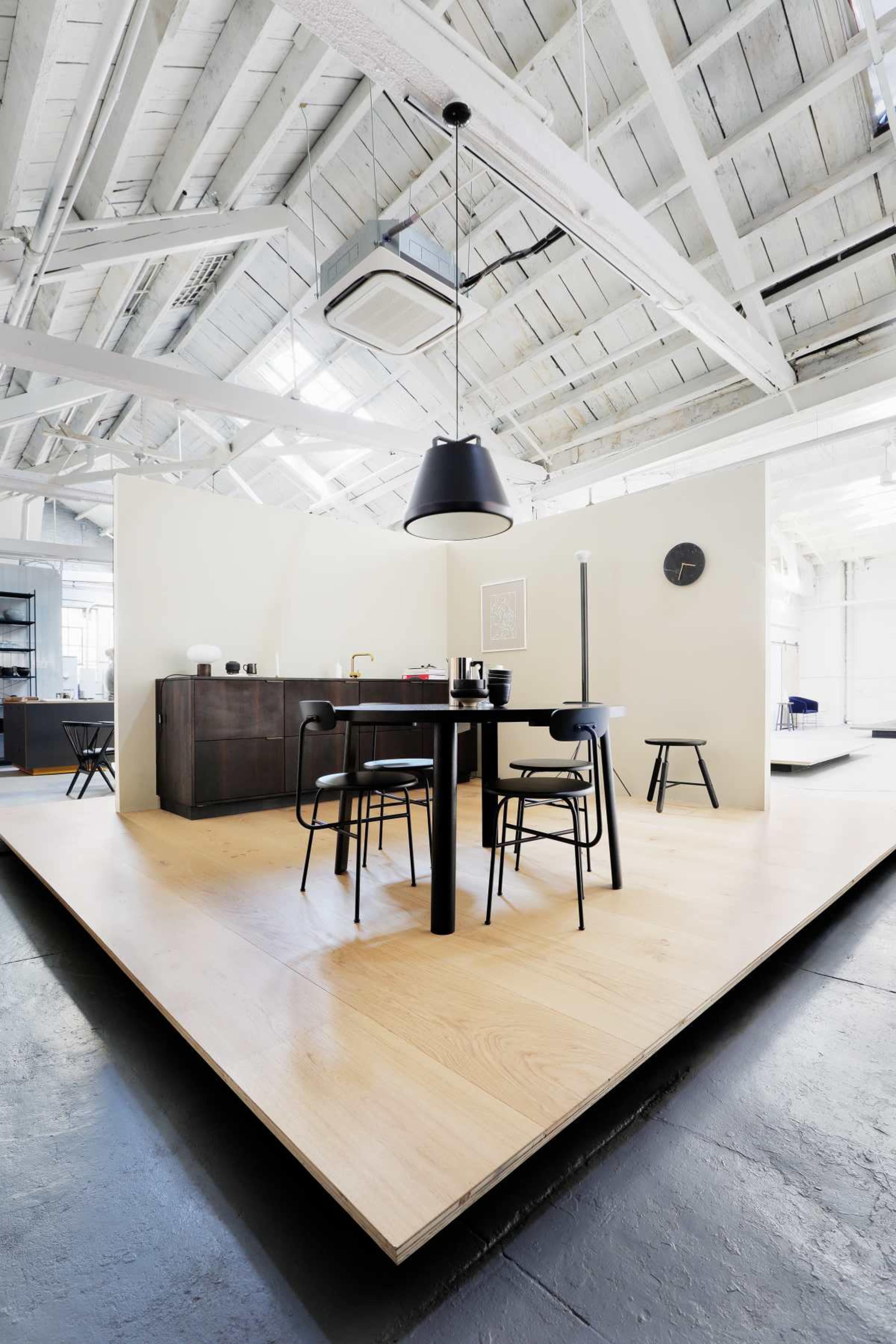 A modern dining area features a black table with chairs, a suspended black light fixture, and a partially enclosed kitchen in an open space with high ceilings and large windows.