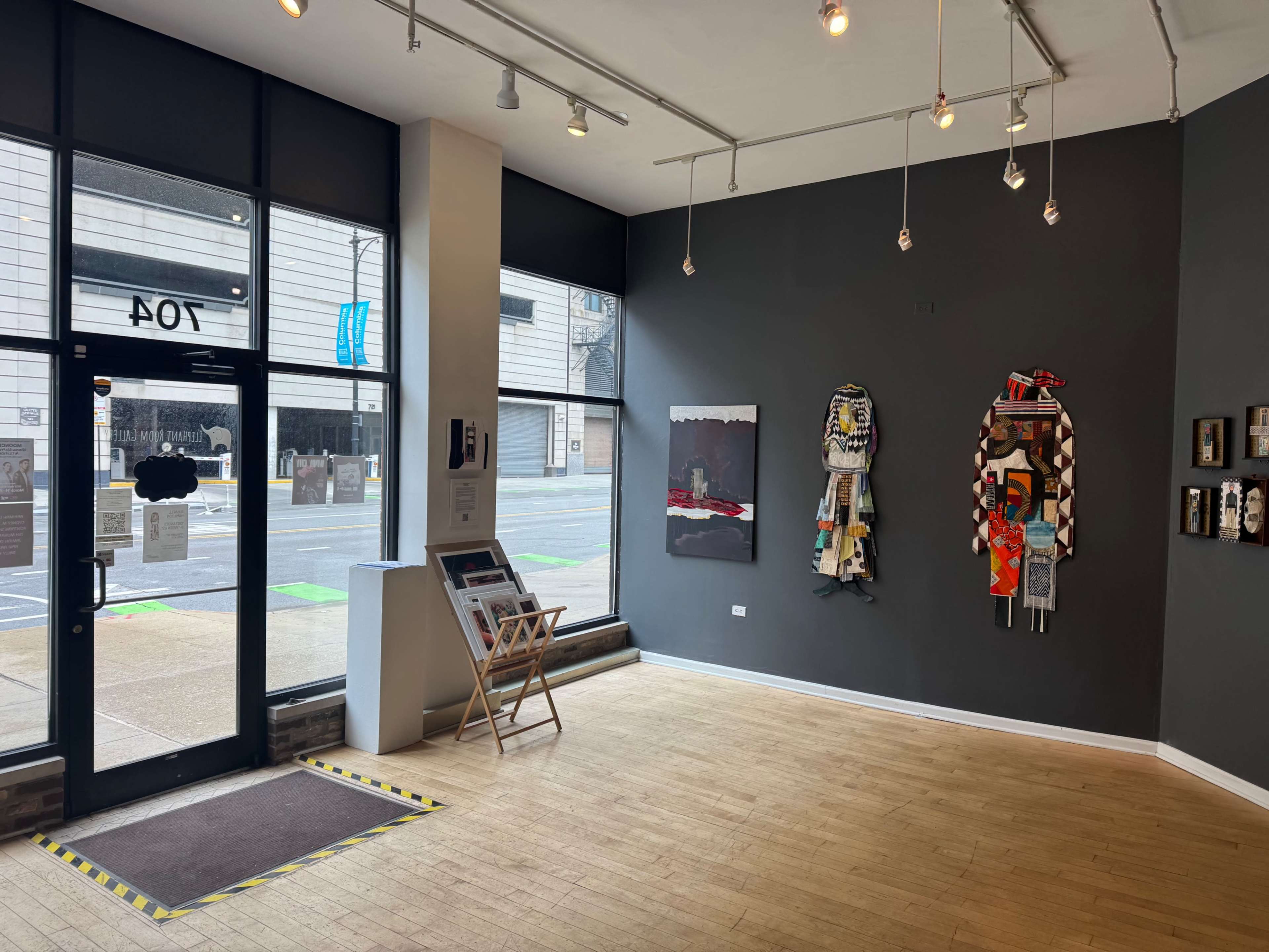 The image shows the interior of an art gallery with two artworks displayed on a dark wall and a front entrance featuring large glass windows.