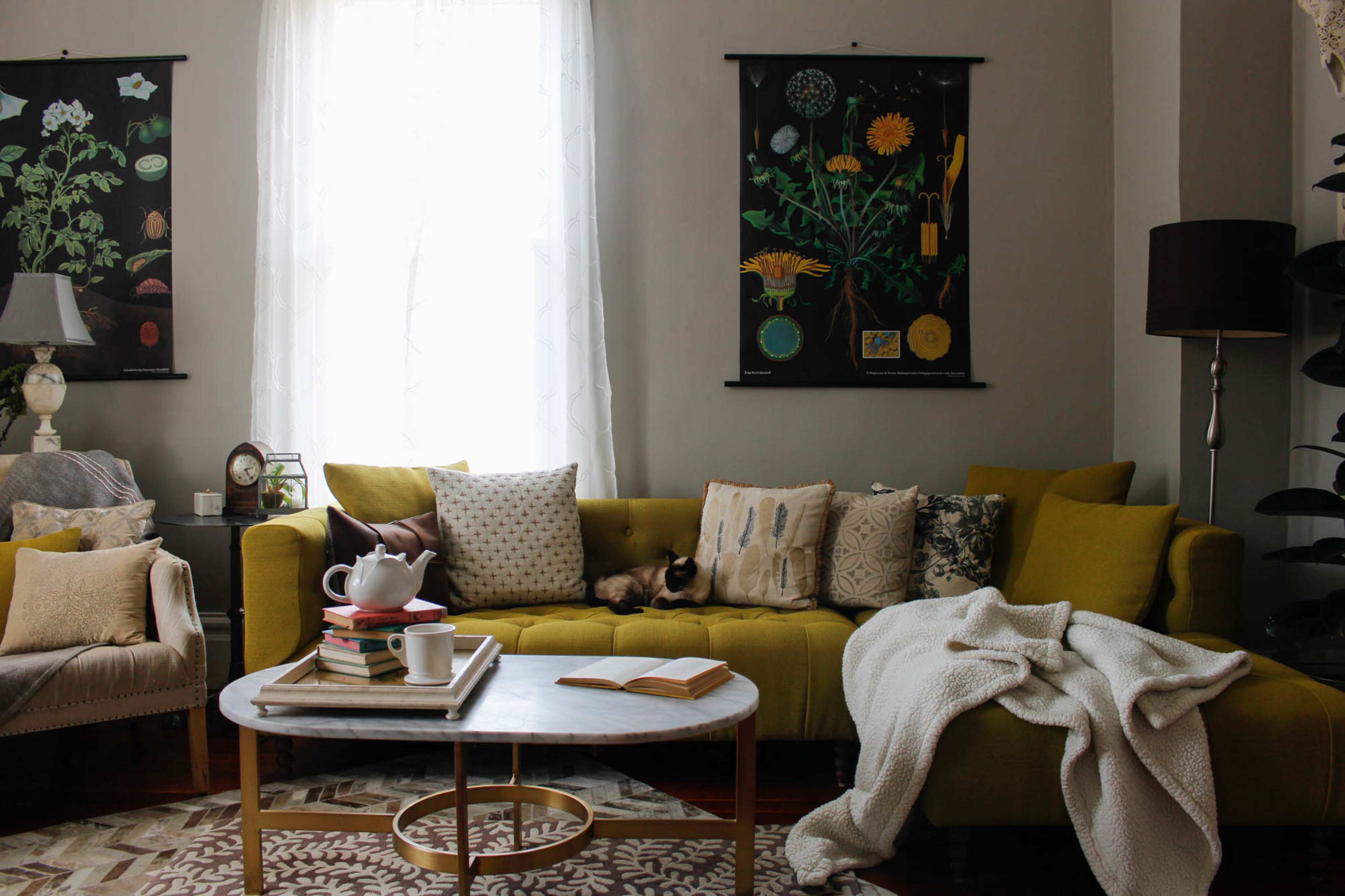A cozy living room features a yellow sofa adorned with decorative pillows, a marble coffee table with books and a teapot, and wall hangings showcasing botanical prints.