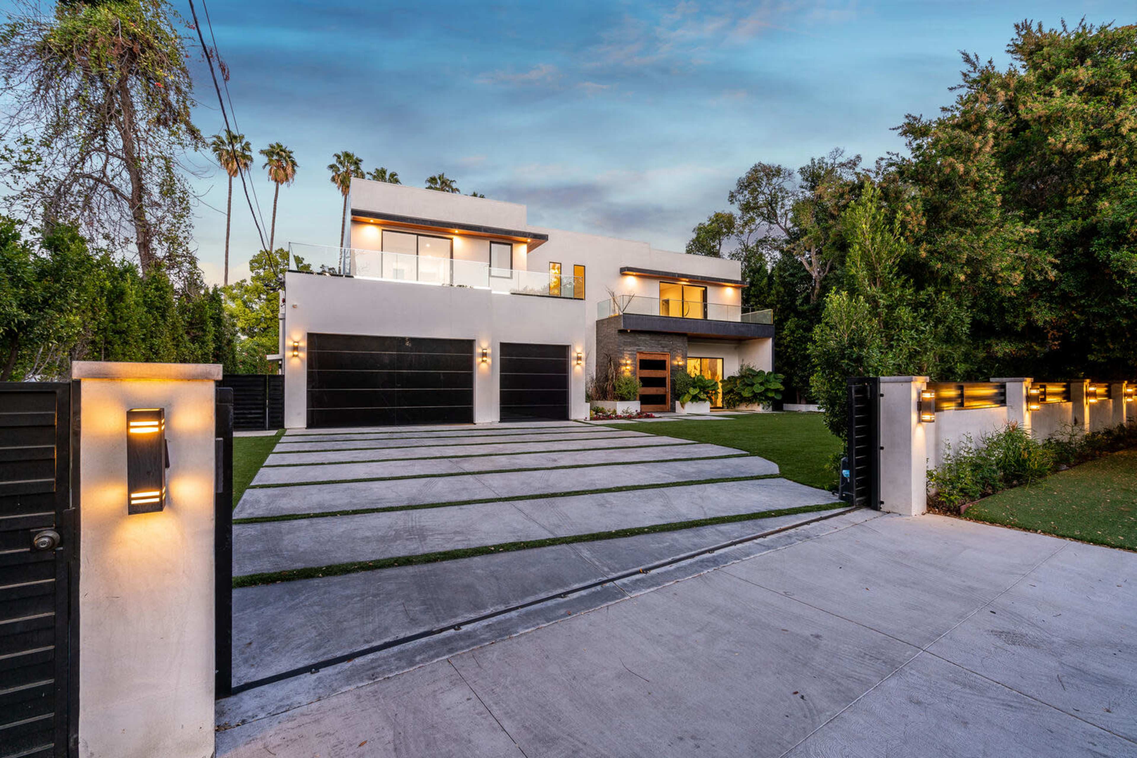 A modern, two-story house with a manicured lawn and a wide driveway is set behind a black gate.