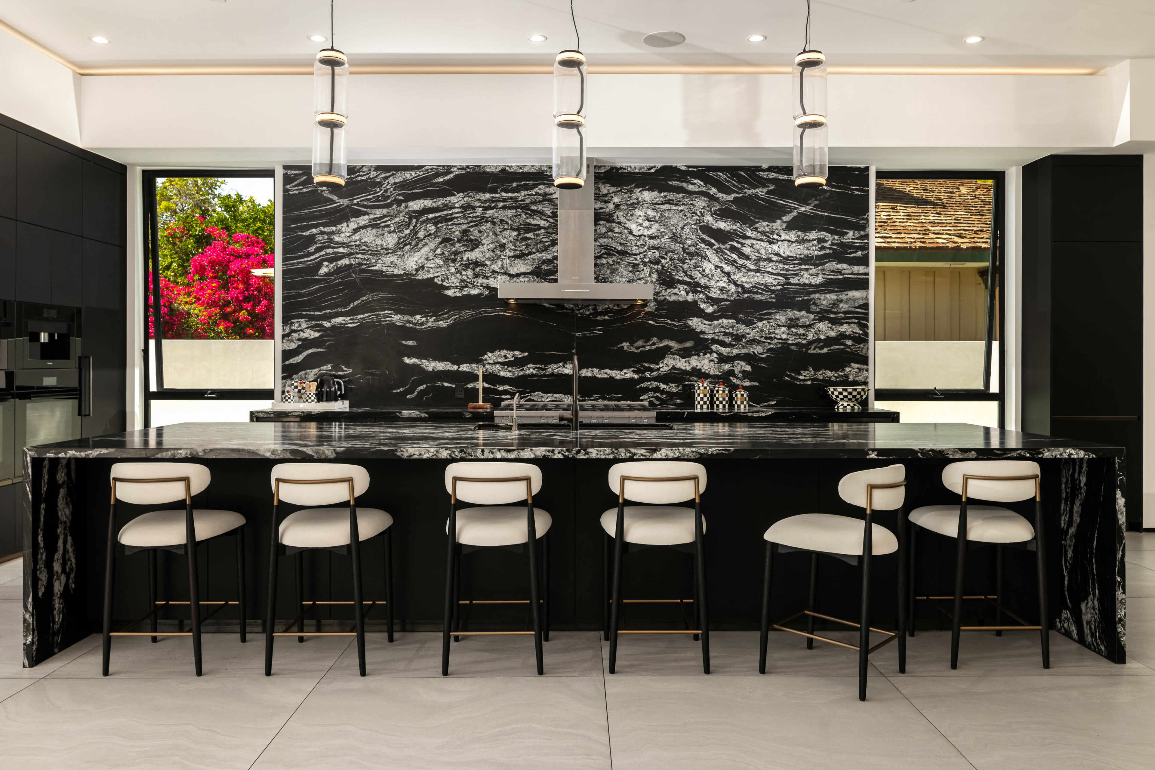 The image shows a modern kitchen with a black marble countertop, high-backed stools, and large windows featuring vibrant flowers outside.