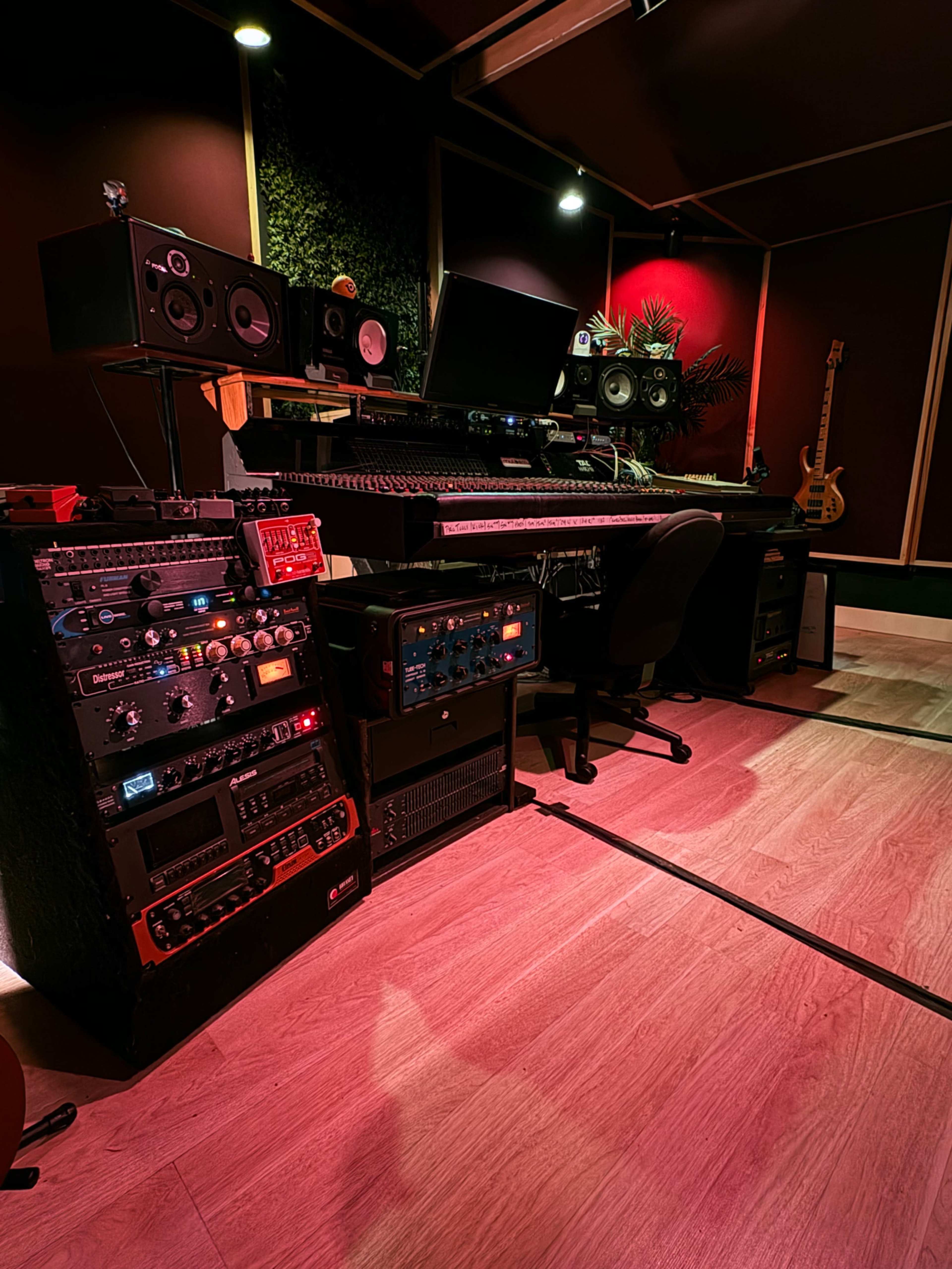 The image shows a modern music studio featuring a large mixing console, various equipment racks with illuminated controls, and sound monitors against a backdrop of red and green lighting.
