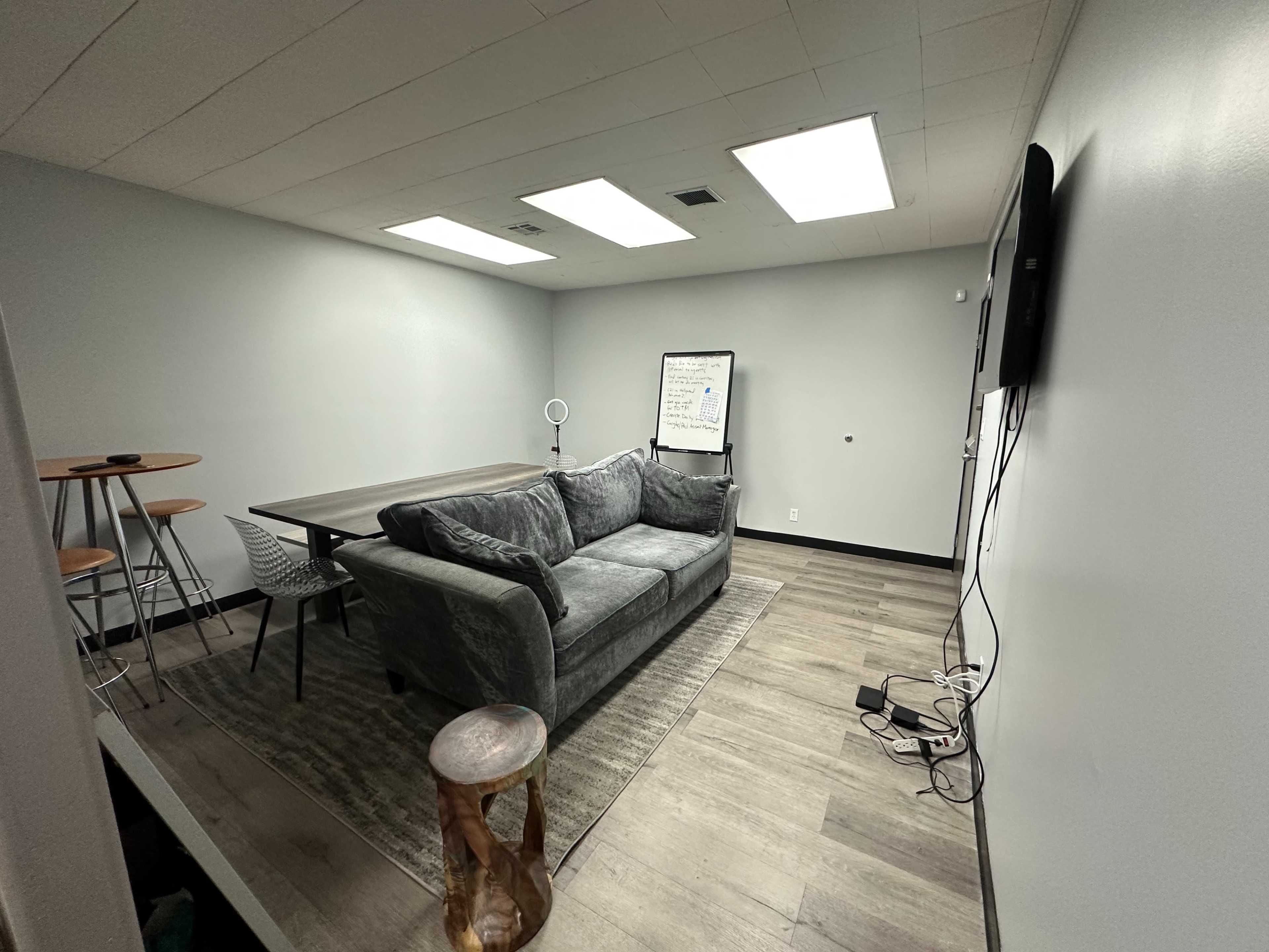 A gray seating area with a couch, a table with stools, and a whiteboard in a minimally furnished room.