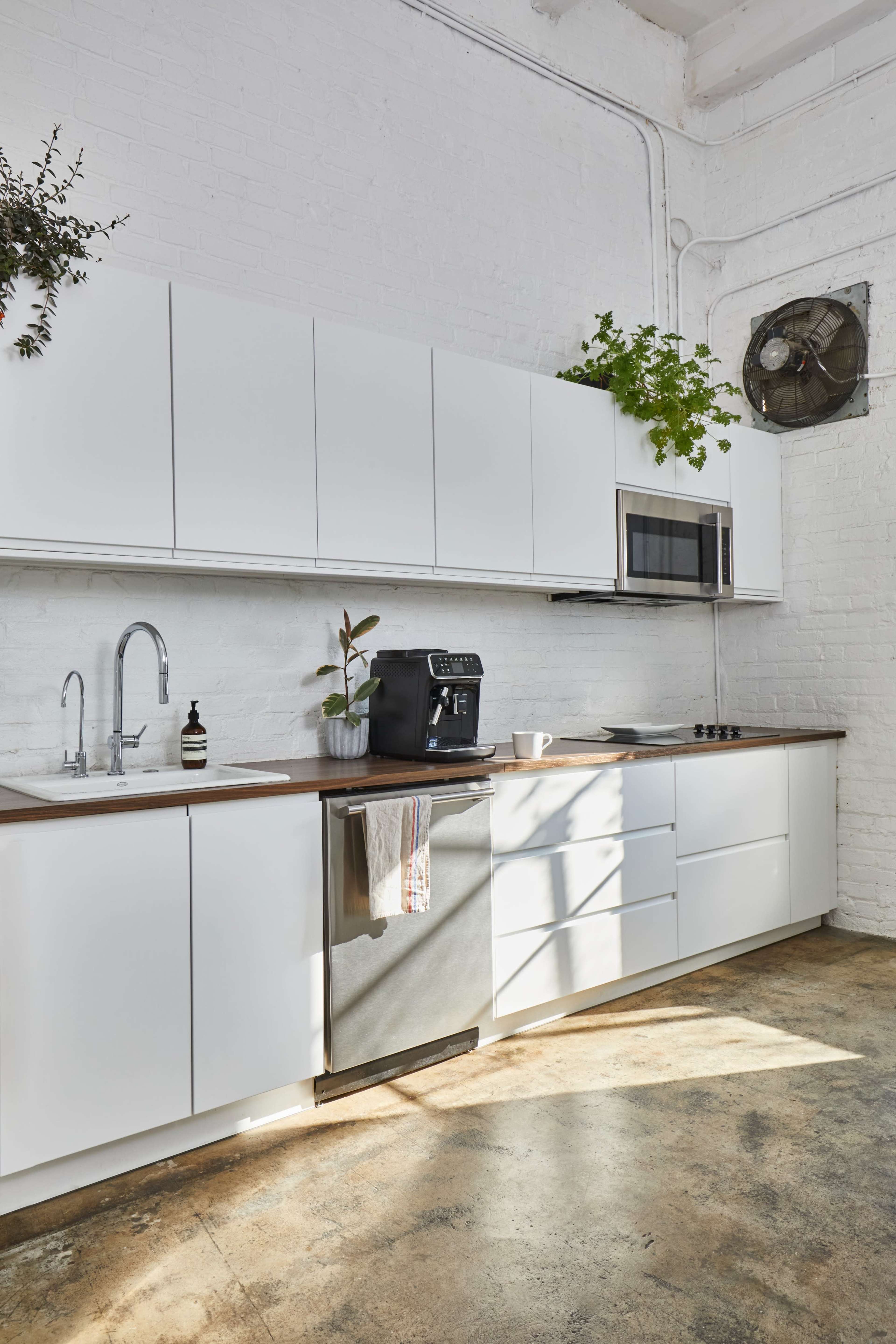 A modern kitchen with white cabinetry, a stainless steel refrigerator, a coffee machine, and plants, illuminated by natural light from a nearby window.
