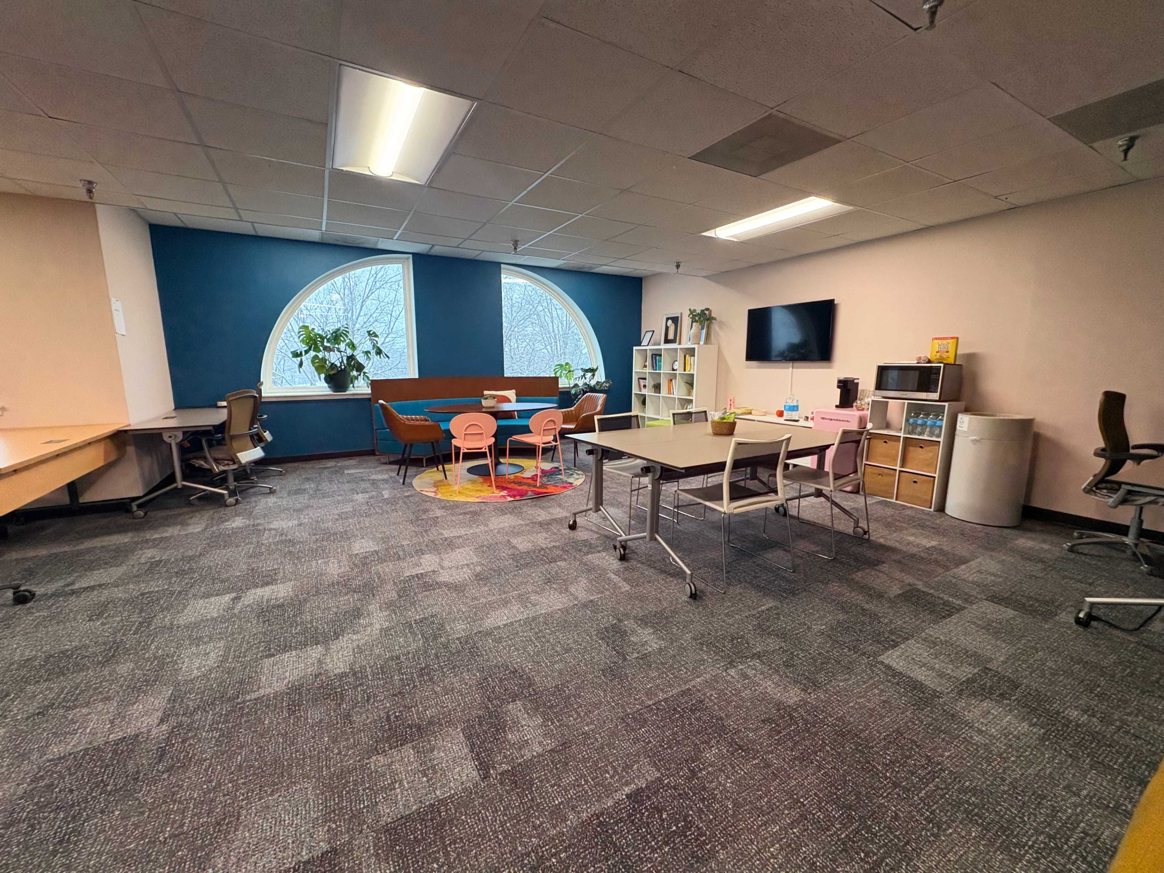 The image shows a modern office space with desks, chairs, and a sitting area featuring a large window and plants.