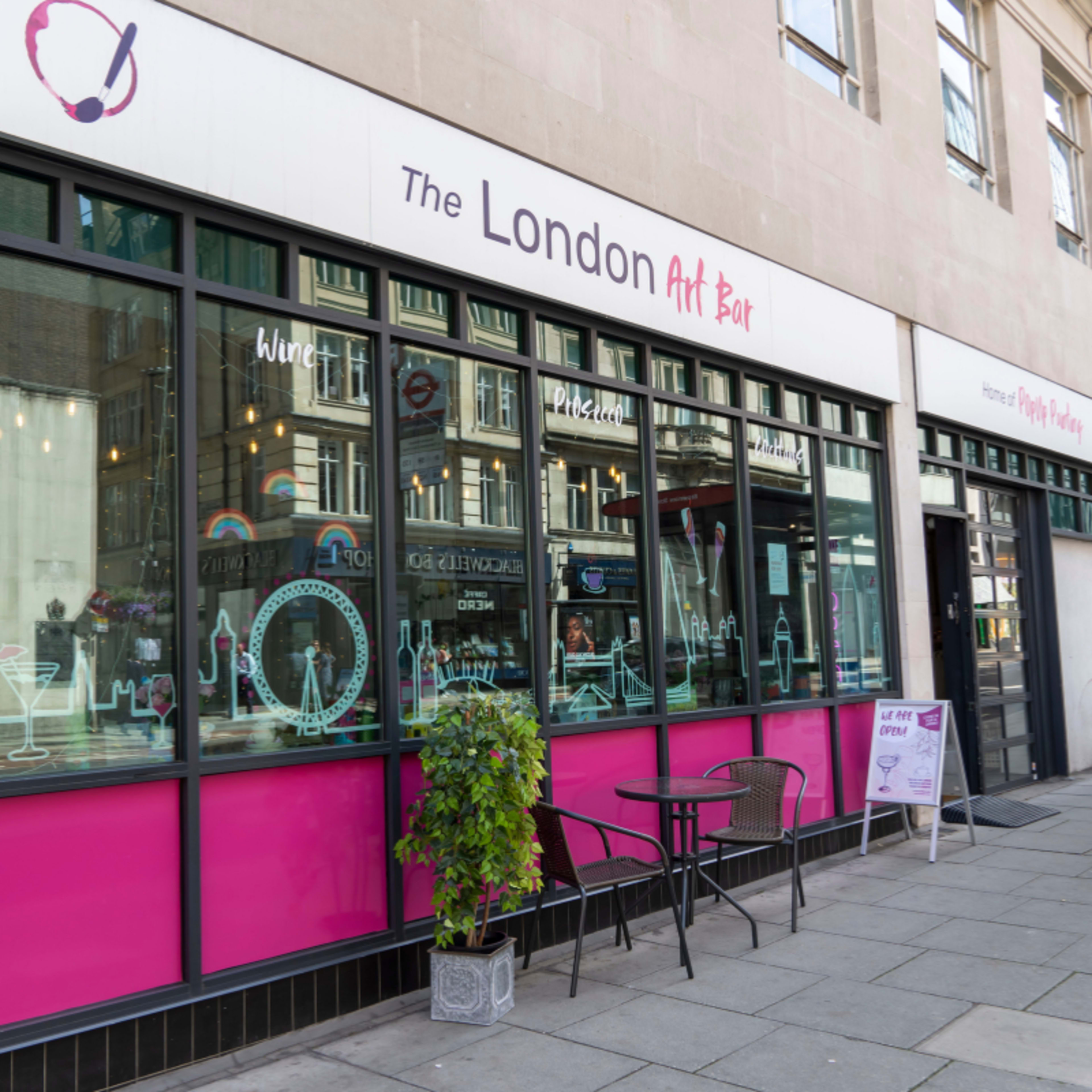 The image shows a storefront labeled "The London Art Bar," featuring large windows with a vibrant pink border, outdoor seating, and a sign advertising various drinks.