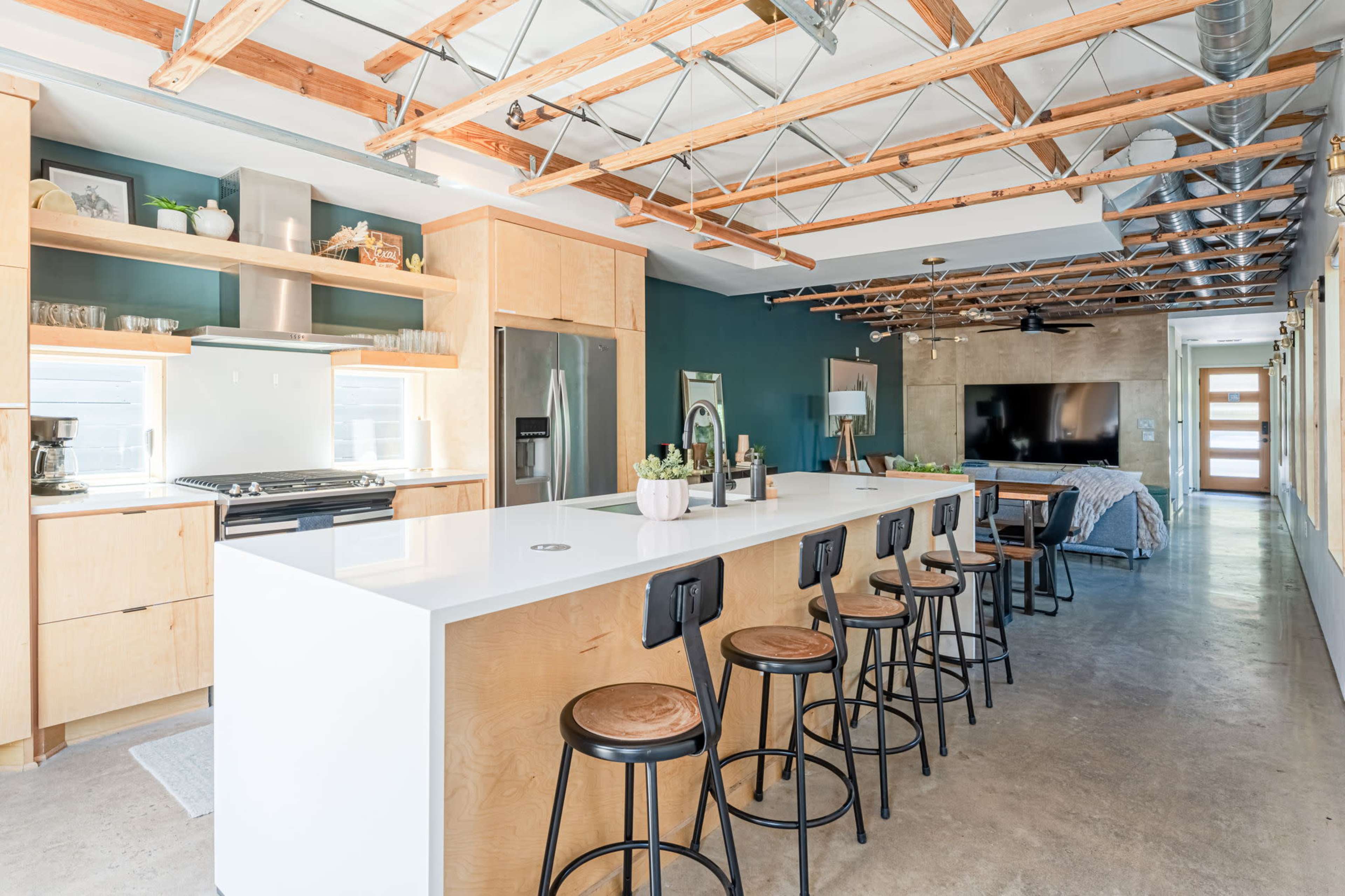 The image shows a modern kitchen and dining area with a large white island, wooden cabinetry, and exposed beams on the ceiling.