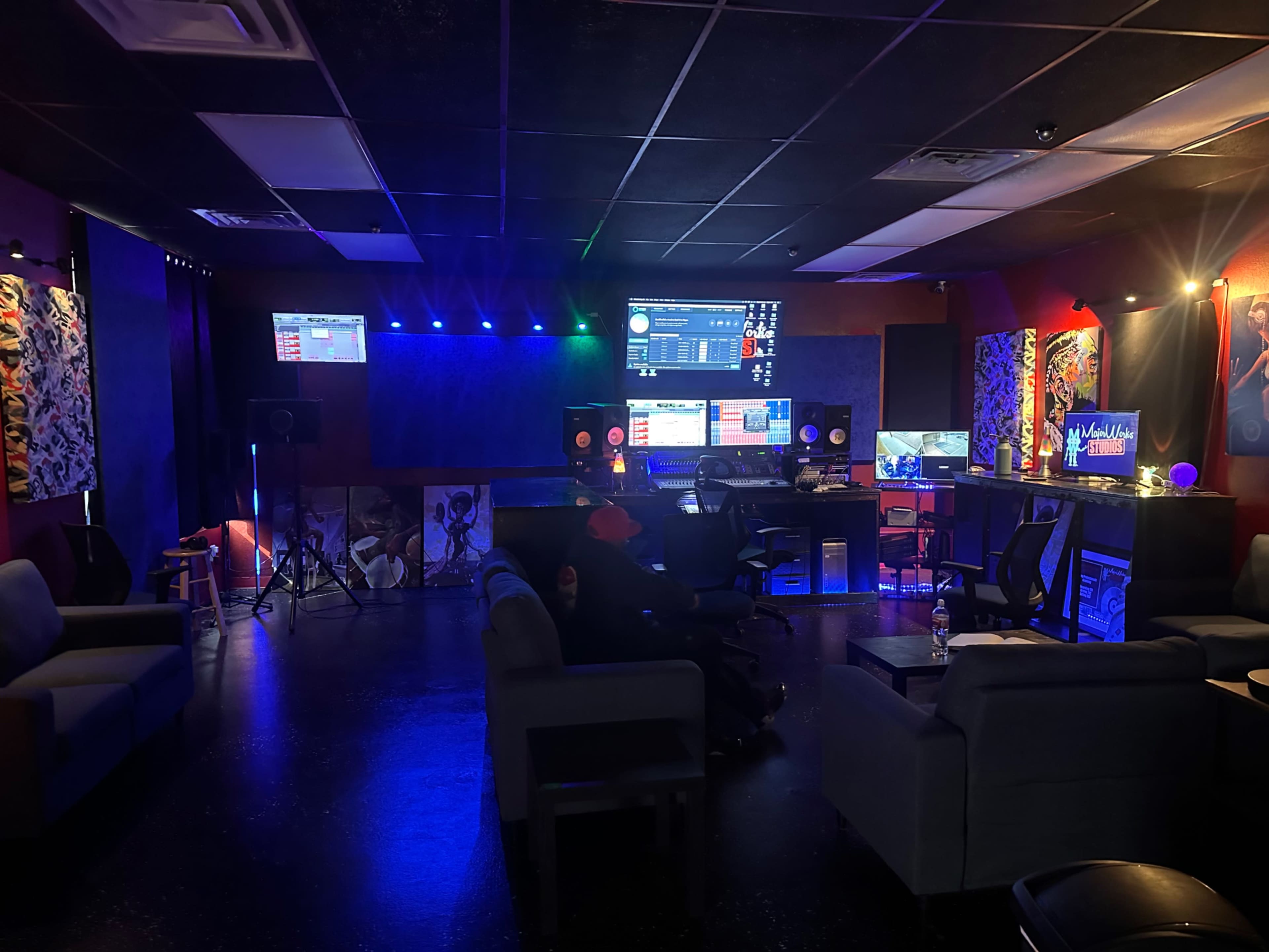 The image shows a music studio with modern equipment, dim lighting, and seating areas scattered throughout the room.