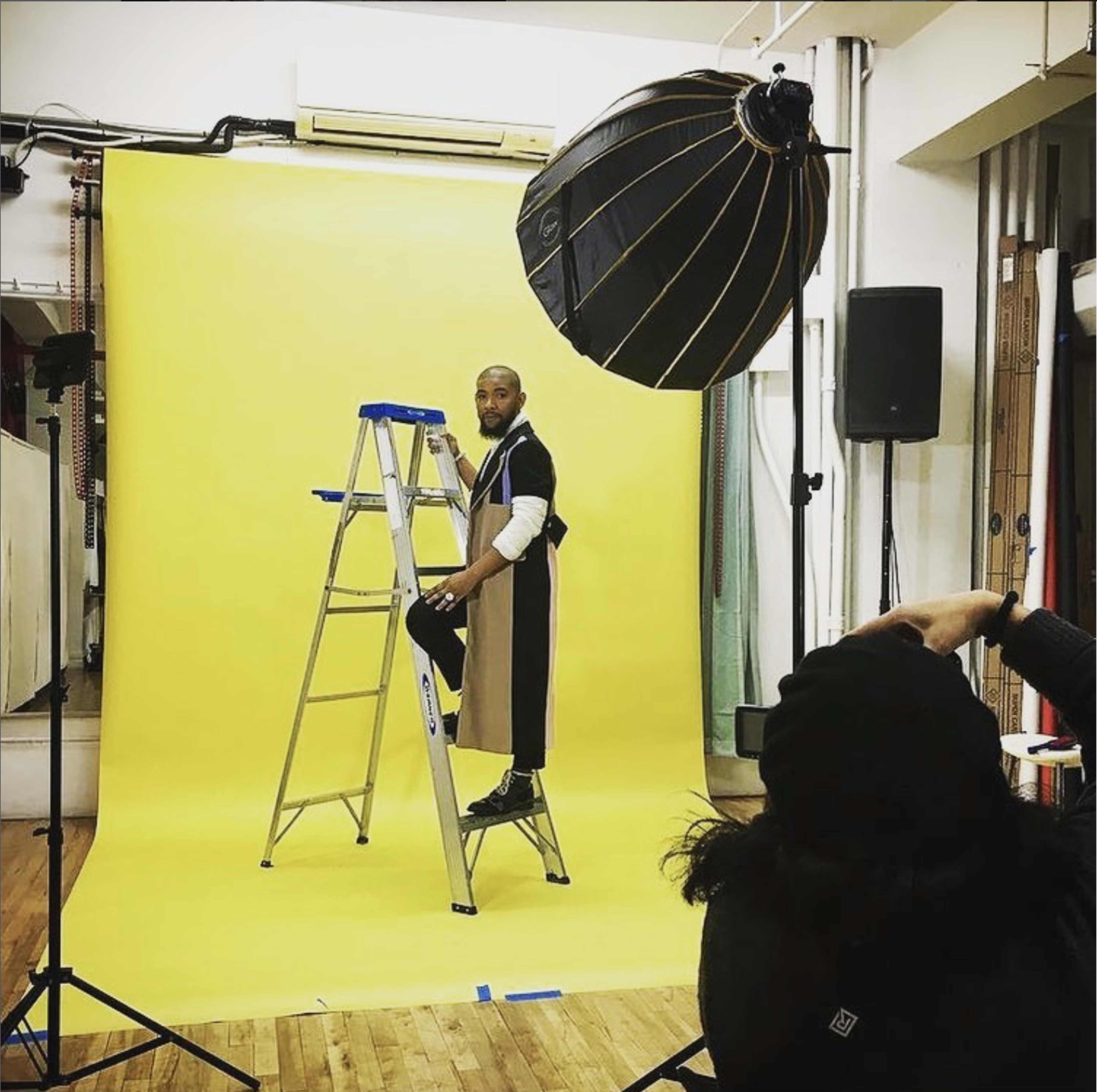 A person stands on a ladder against a yellow backdrop in a photography studio, with a large light source suspended overhead.