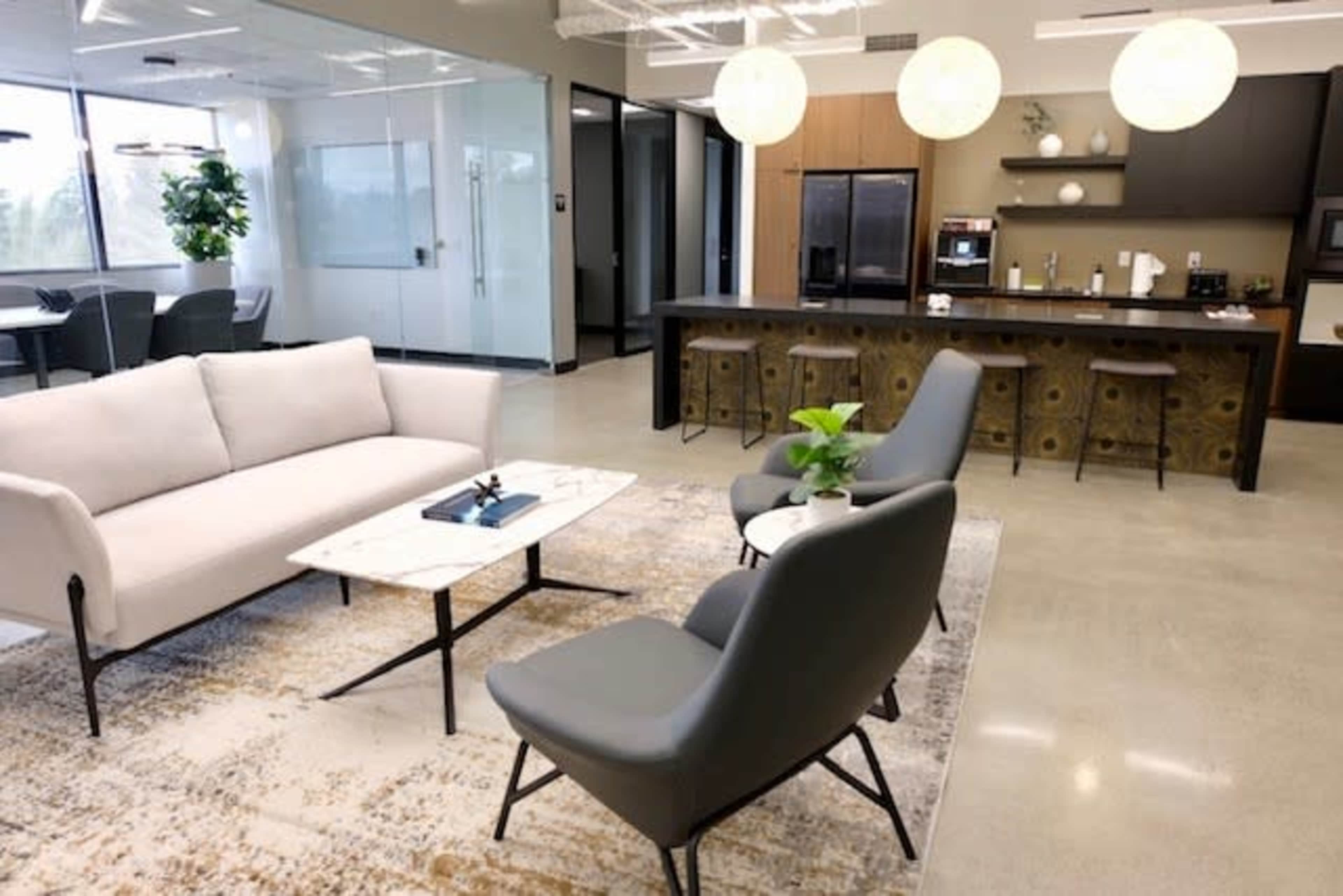 A modern office lounge area with a sofa, two chairs, a coffee table, and a kitchenette in the background.