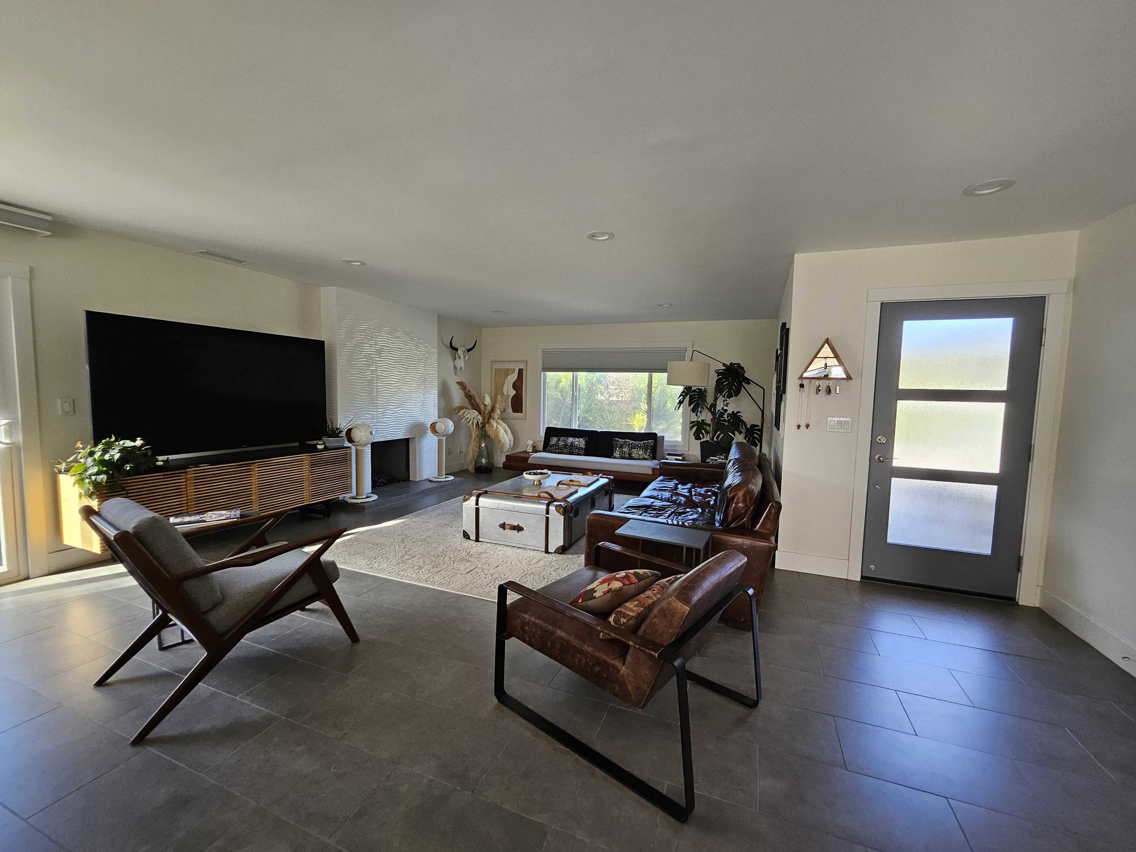 A modern living room featuring a large television, a coffee table, two armchairs, and a door leading outside.