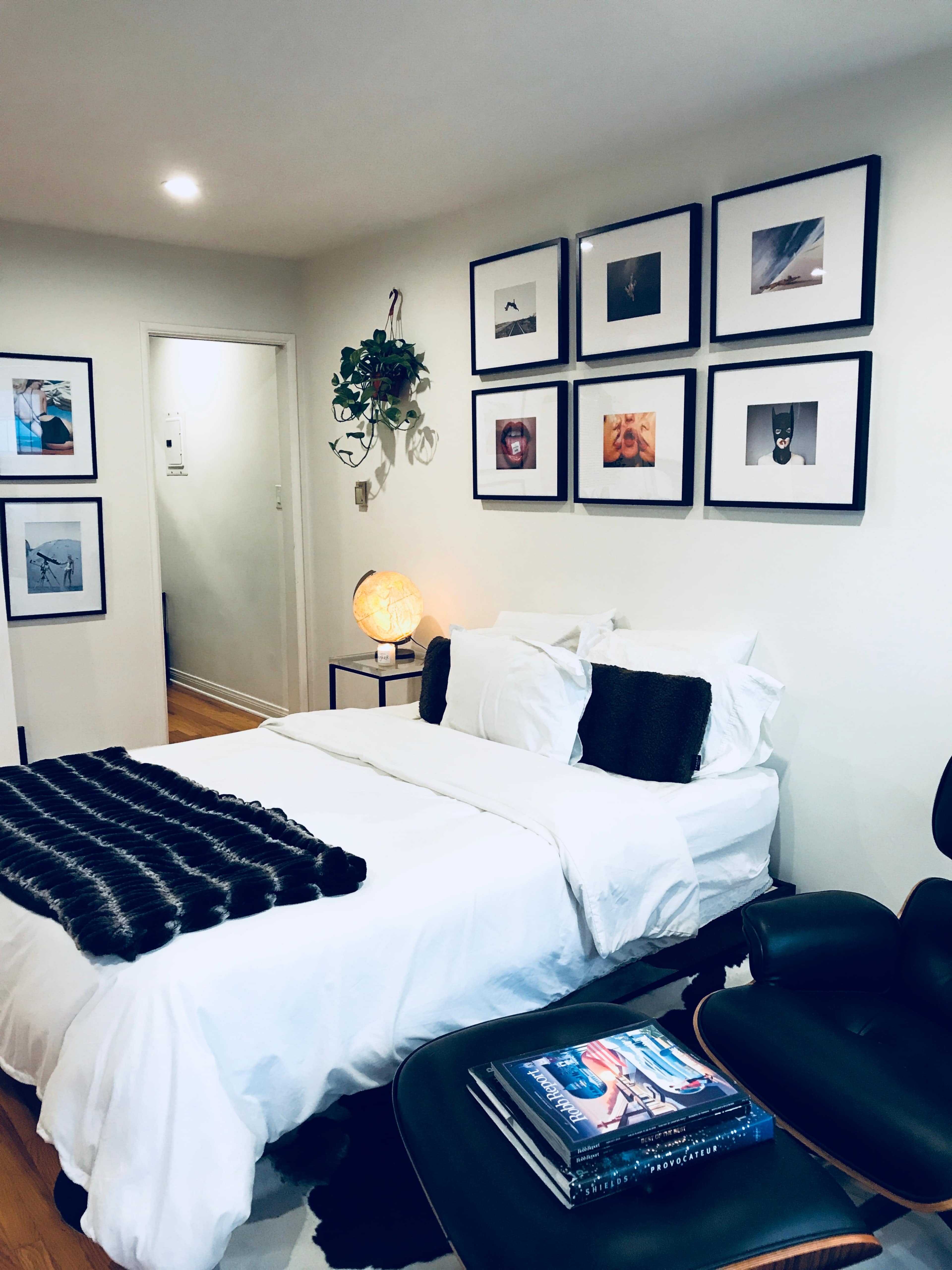 A neatly arranged bedroom features a bed with white linens, a dark throw blanket, framed photographs on the wall, and a small side table with books.