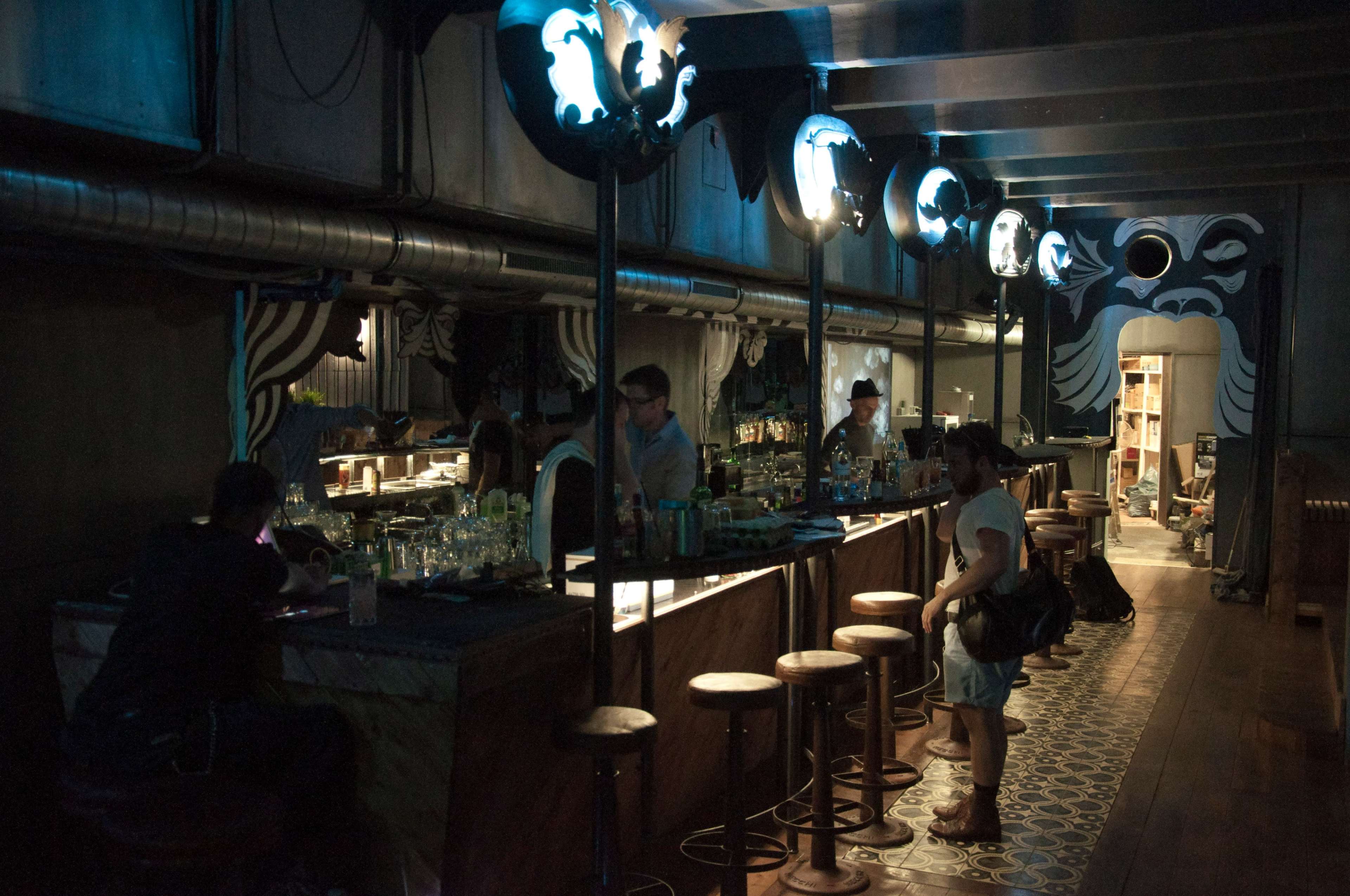 A dimly lit bar features wooden stools and illuminated circular designs on the walls while patrons engage in conversation.